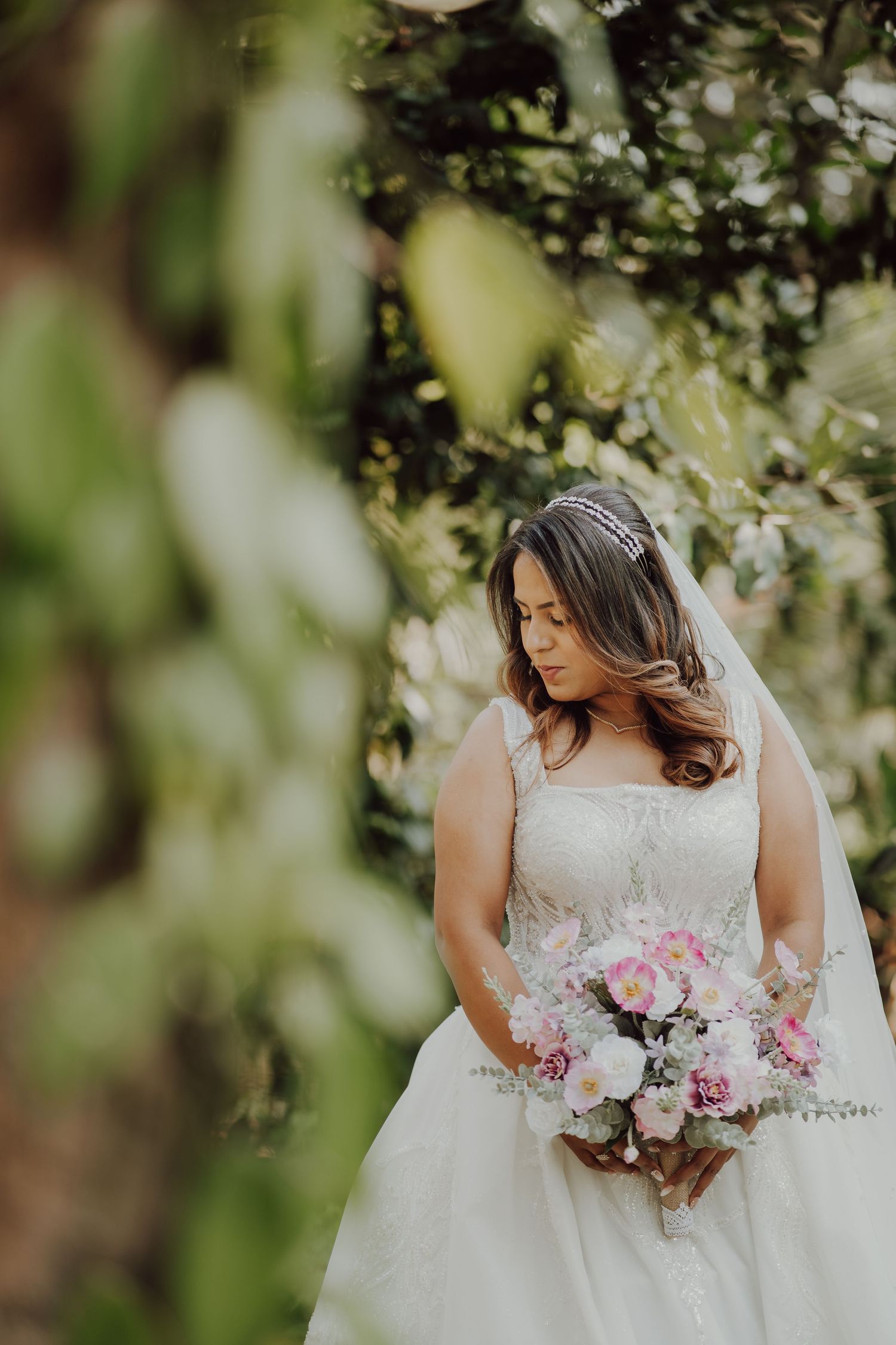 A bride in a white lace gown holds a pink and white bouquet in a garden setting.