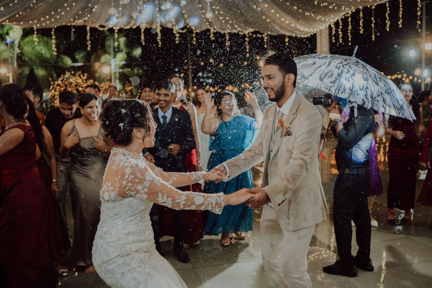 Bride and groom share a joyful dance under string lights while a guest holds a clear umbrella over the festive outdoor reception.