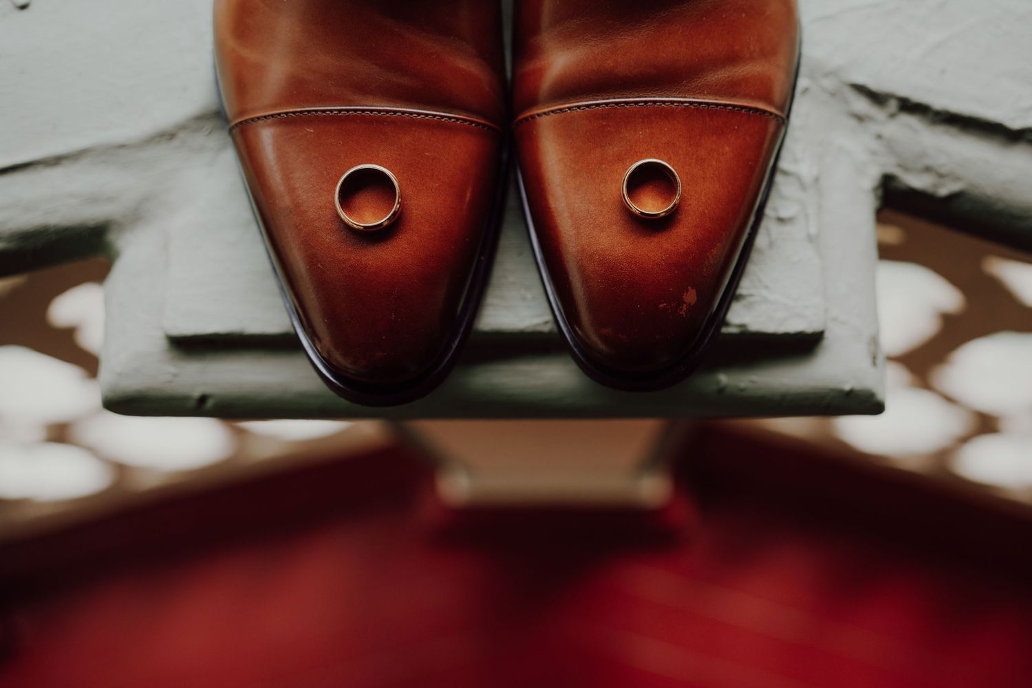 Two wedding rings rest on the toes of polished brown leather cap-toe dress shoes placed on a stone ledge above a red patterned floor.