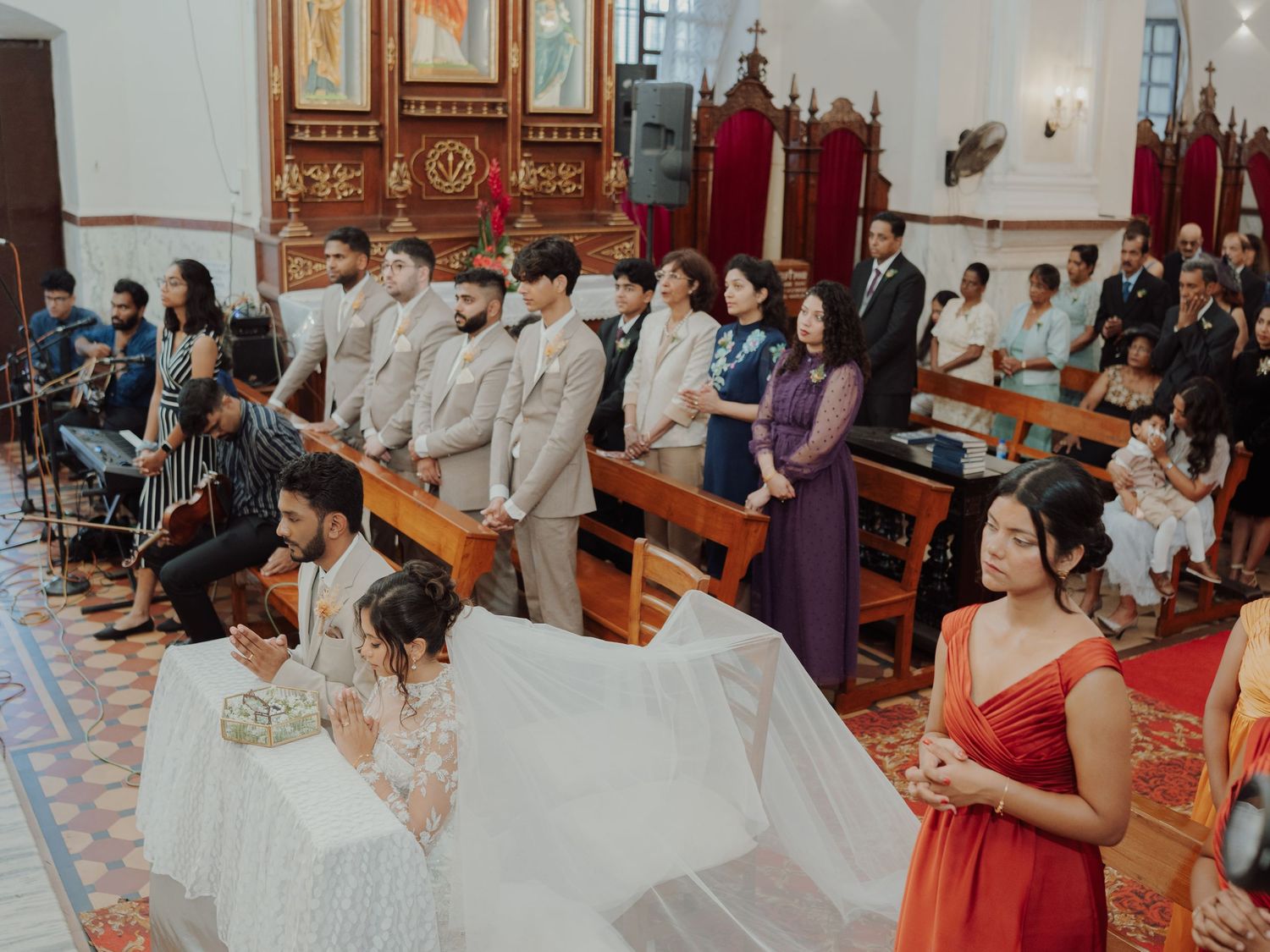 Wedding ceremony inside a traditional church as a couple kneels at the altar while guests and musicians look on from wooden pews.