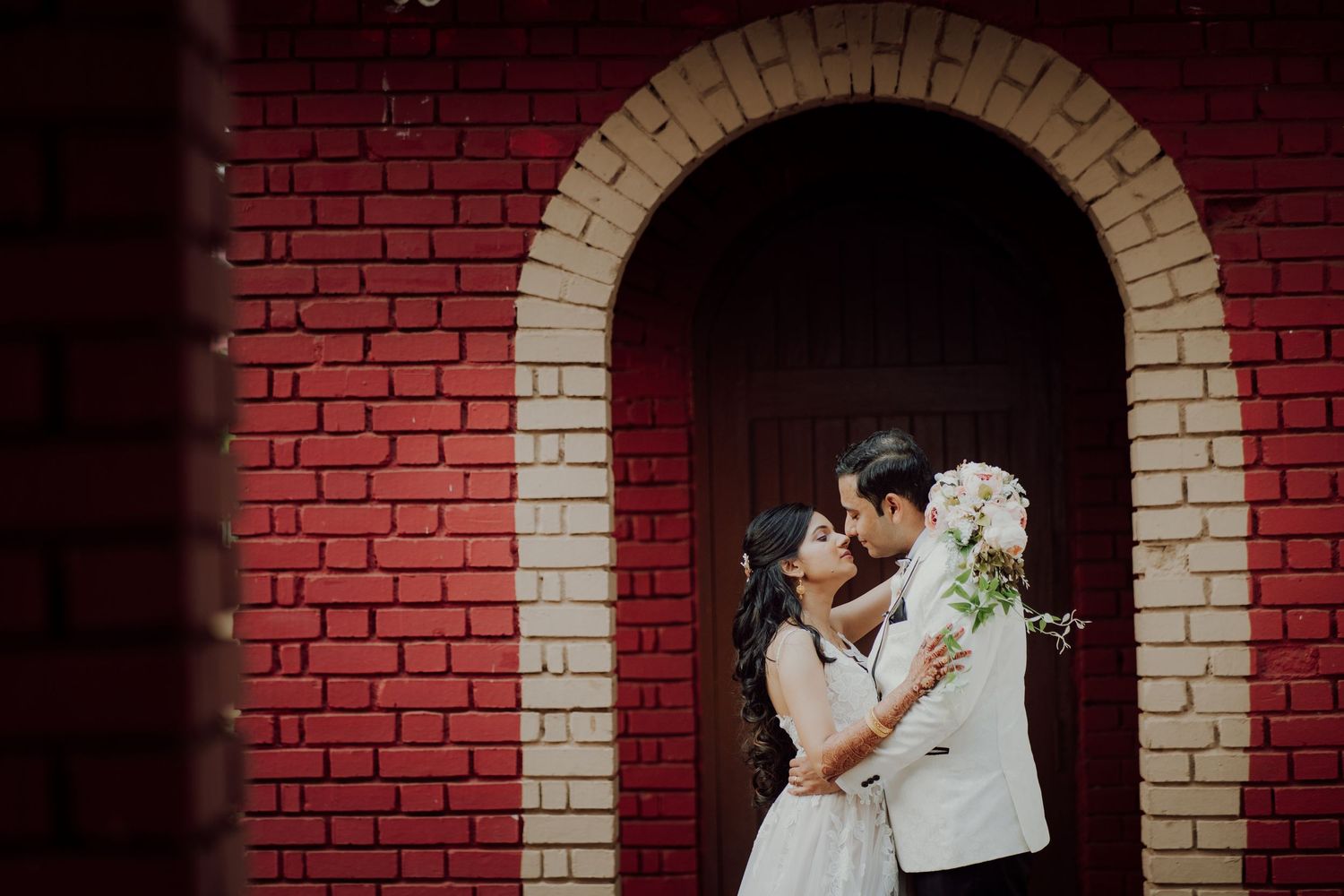 Romantic wedding couple embraces beneath brick archway with white stone trim in dramatic lighting.