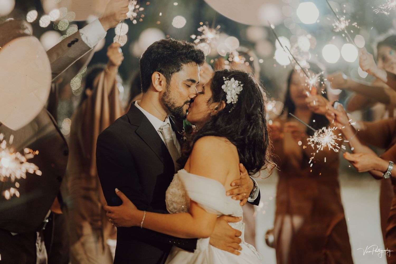 A romantic wedding couple shares an intimate kiss surrounded by sparklers and soft bokeh lights at their evening celebration.