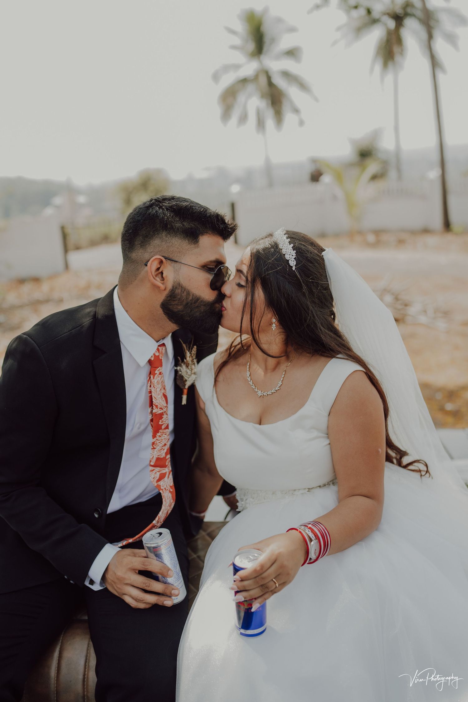 Newlywed couple sharing a romantic moment while holding beverages in a tropical setting with palm trees.