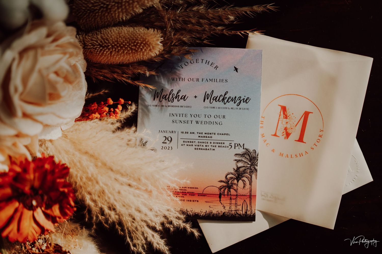 Wedding invitation suite with sunset palm tree design displayed among dried flowers and pampas grass in warm lighting.
