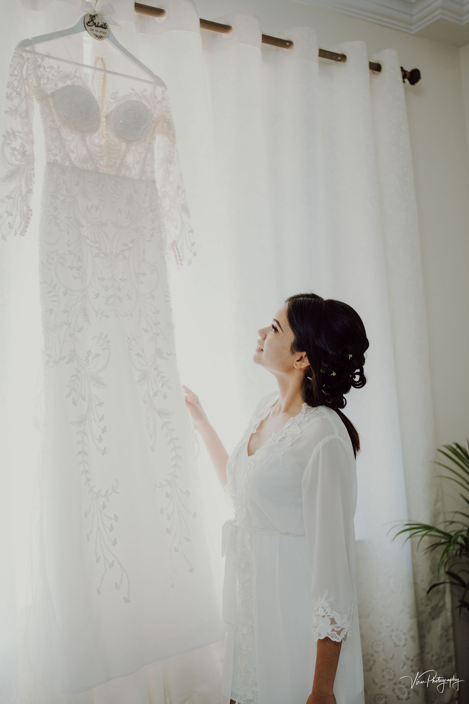A white lace wedding dress hangs on display as a woman in a white robe admires it against a light curtained backdrop.