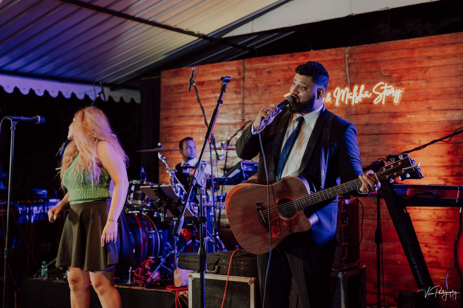 Musicians perform on stage with acoustic guitar at a live music venue with wood panel backdrop and blue lighting.