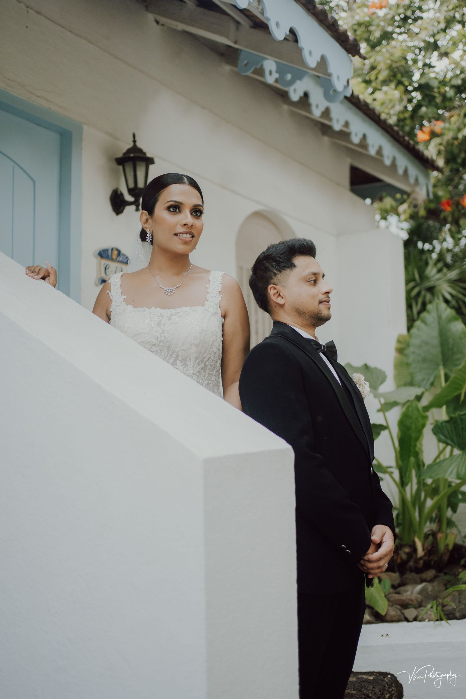 A couple in elegant wedding attire pose on white stairs outside a colonial-style building with turquoise door and greenery.