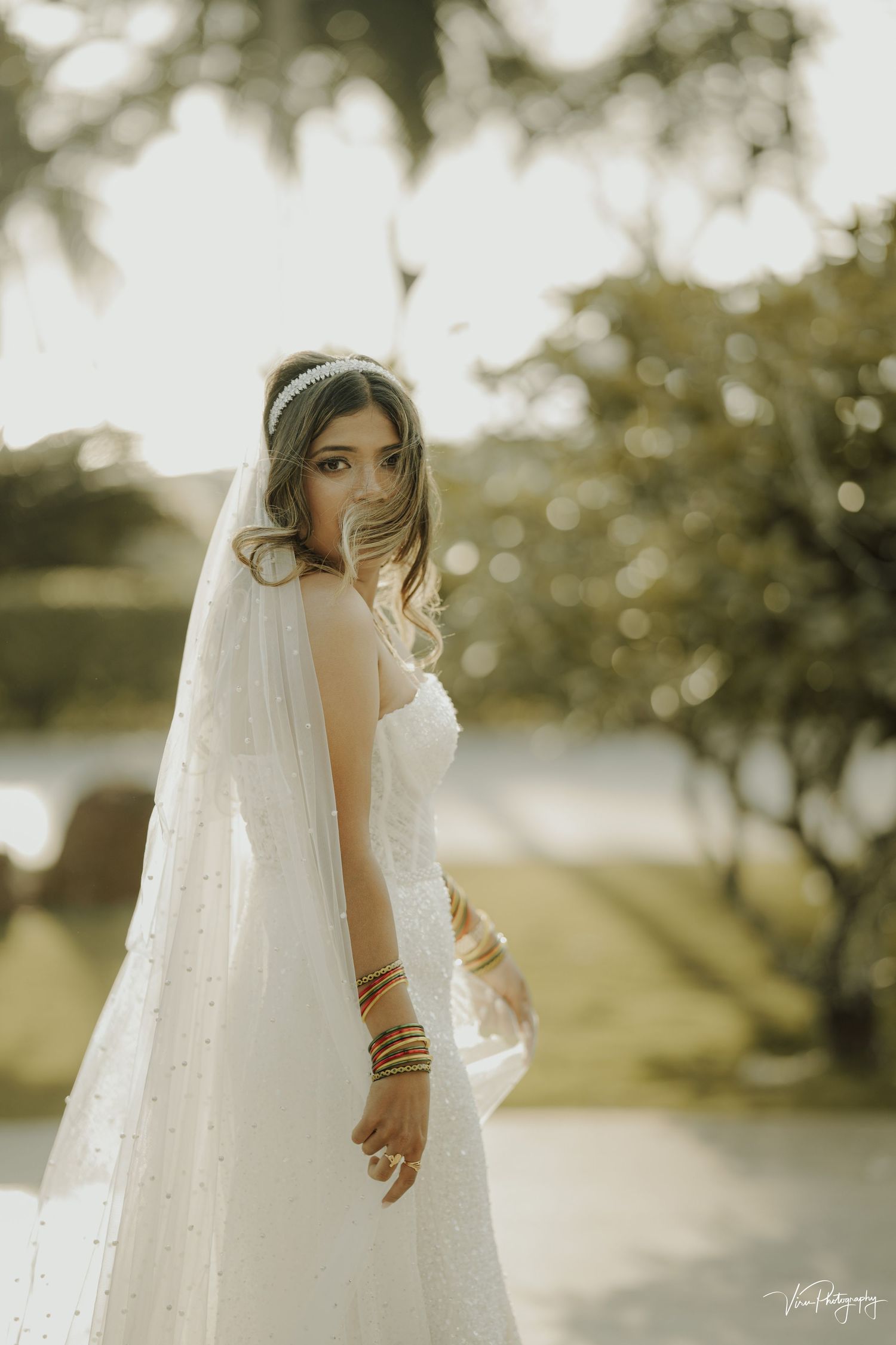 A bride in a flowing white dress with a sparkly veil poses outdoors in dreamy golden hour lighting.