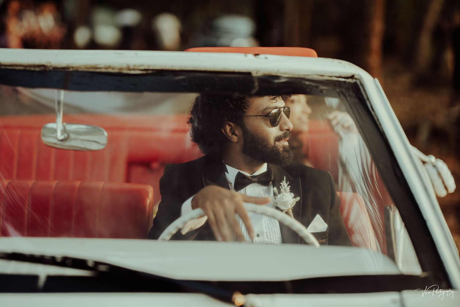 Couple shares a tender moment inside a classic red and white car with palm trees in background.