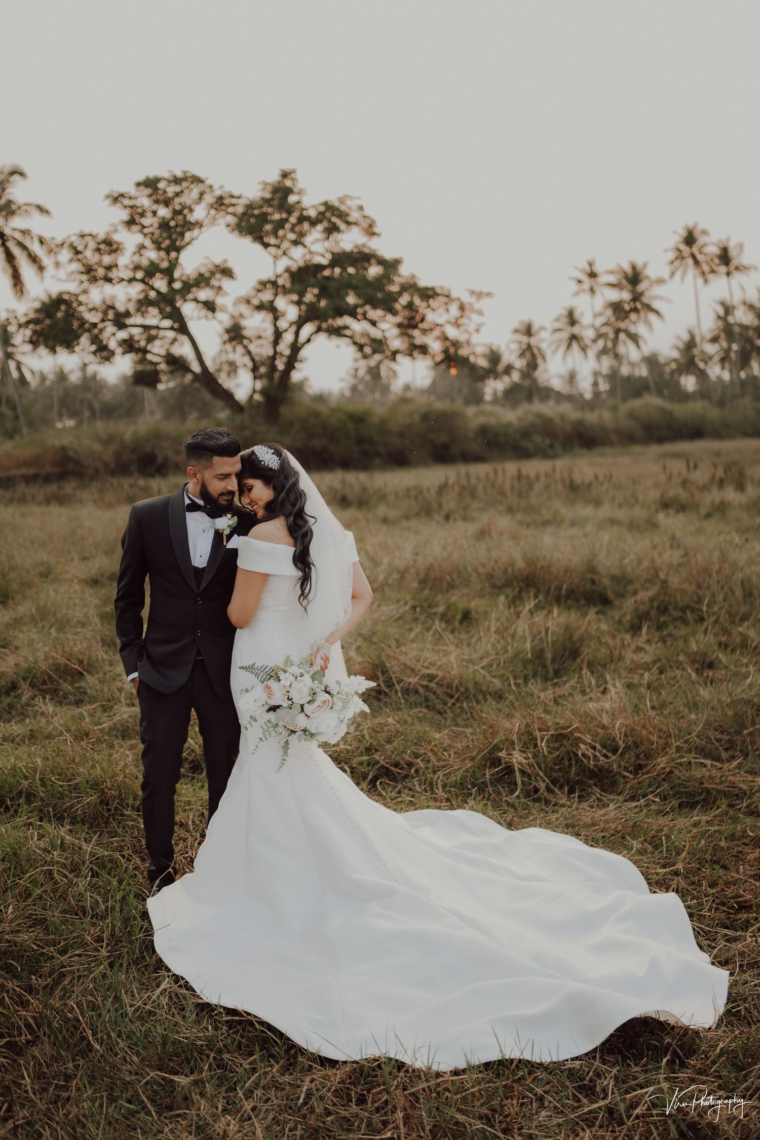 Newlyweds share intimate moment in rustic field with palm trees and flowing white wedding dress at sunset.