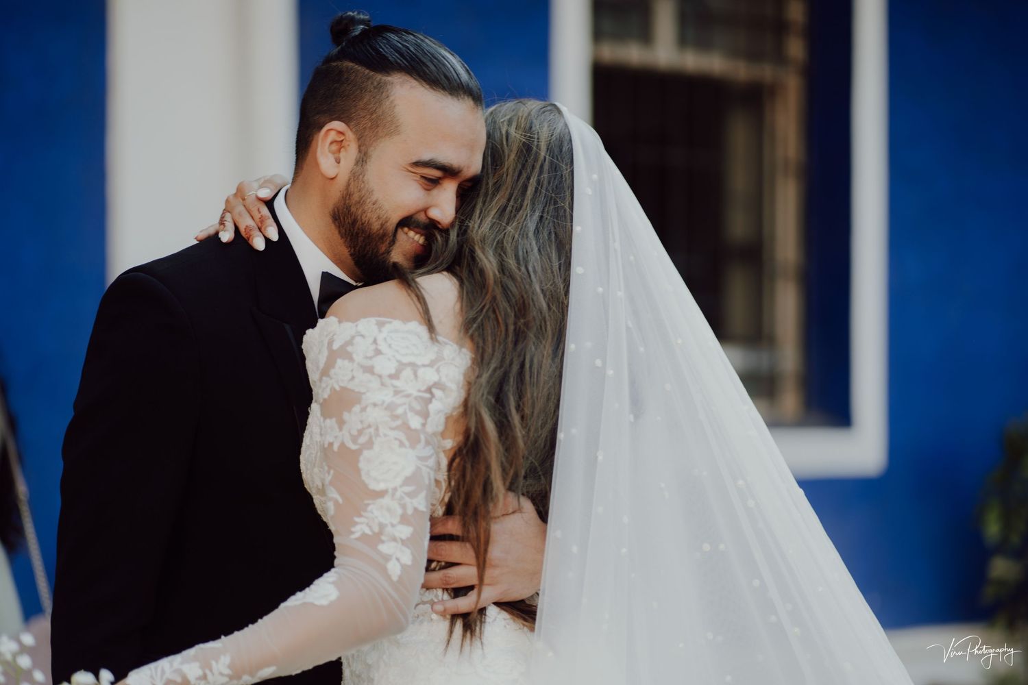 Newlywed couple shares intimate moment together against blue wall with bride wearing lace sleeve wedding dress and flowing veil.