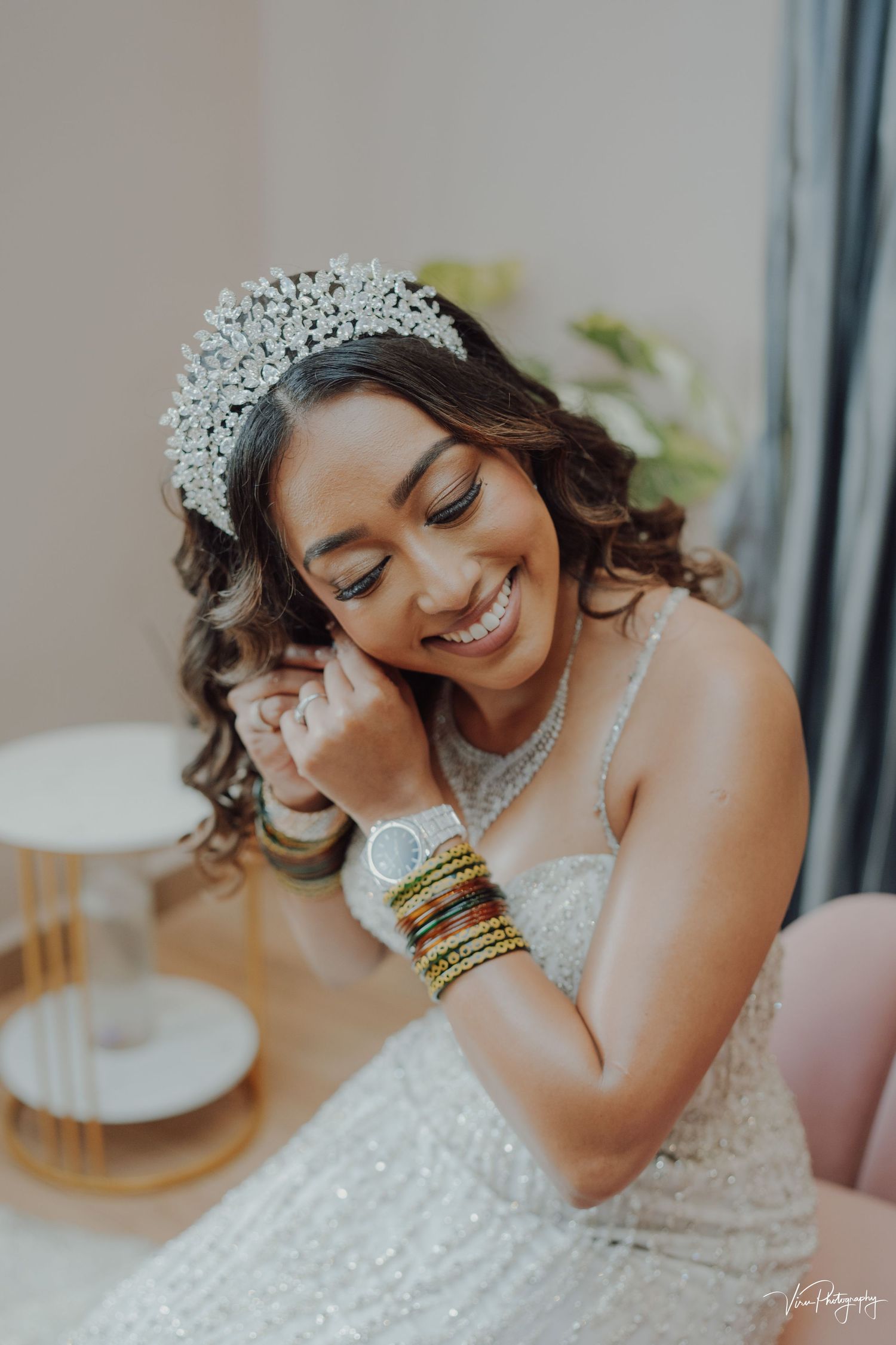Bride wearing sparkly tiara and white dress touches jewelry while smiling with closed eyes in soft natural light.