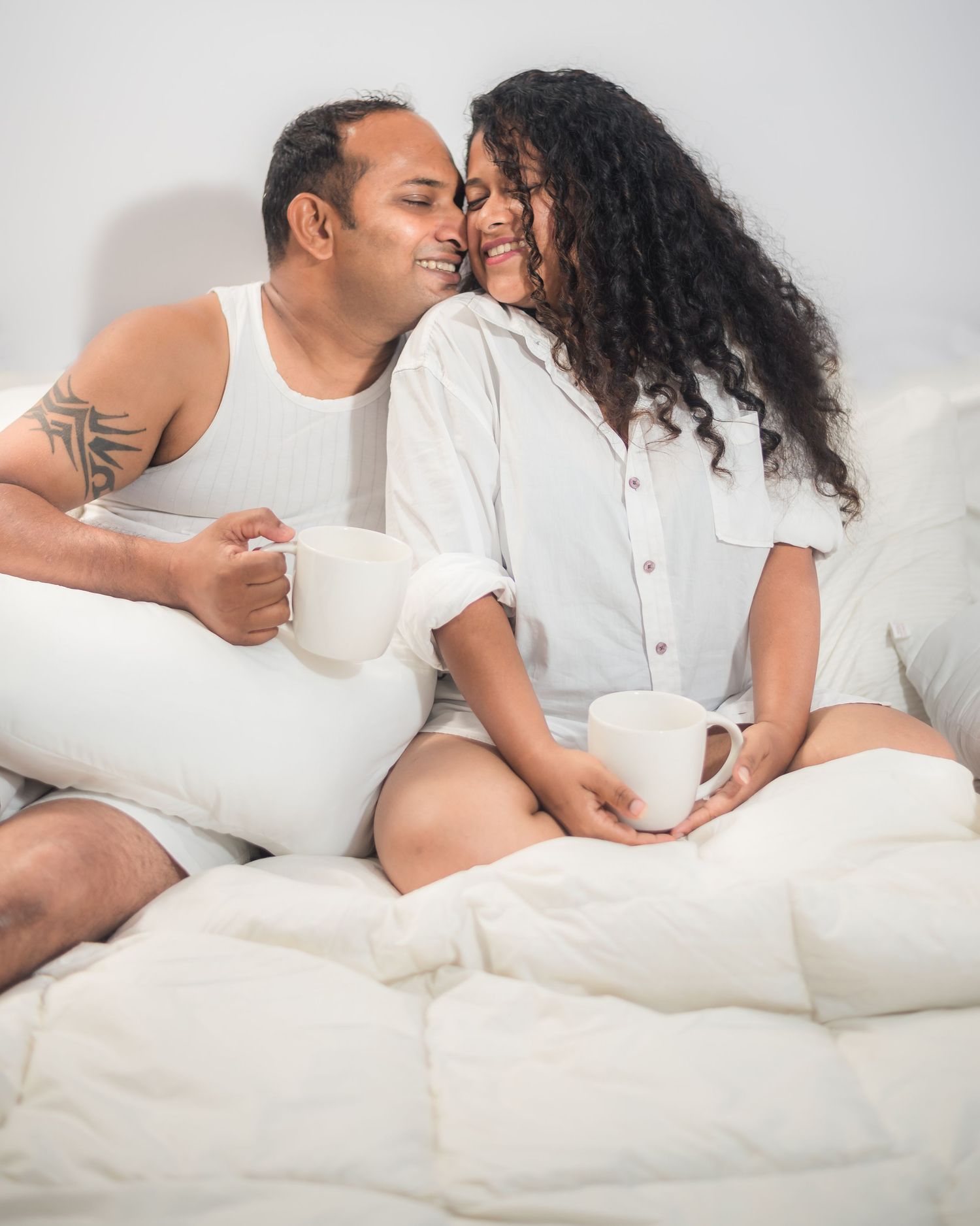 A couple enjoys a cozy morning in bed with coffee cups while sharing affectionate moments.