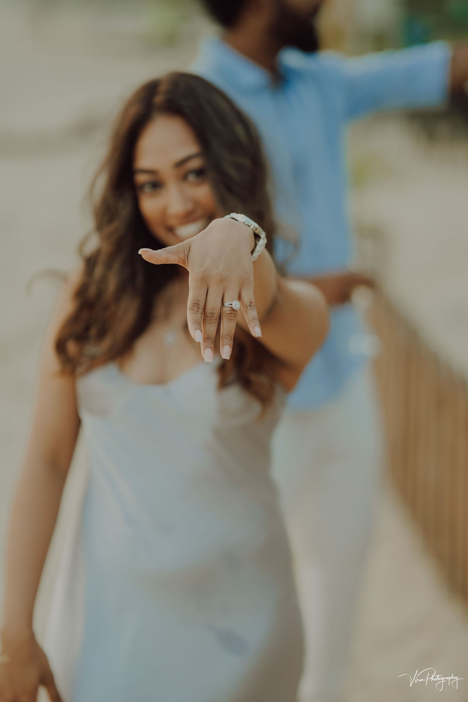 A person wearing a white dress holds out their hand showing an engagement ring in a romantic outdoor setting.