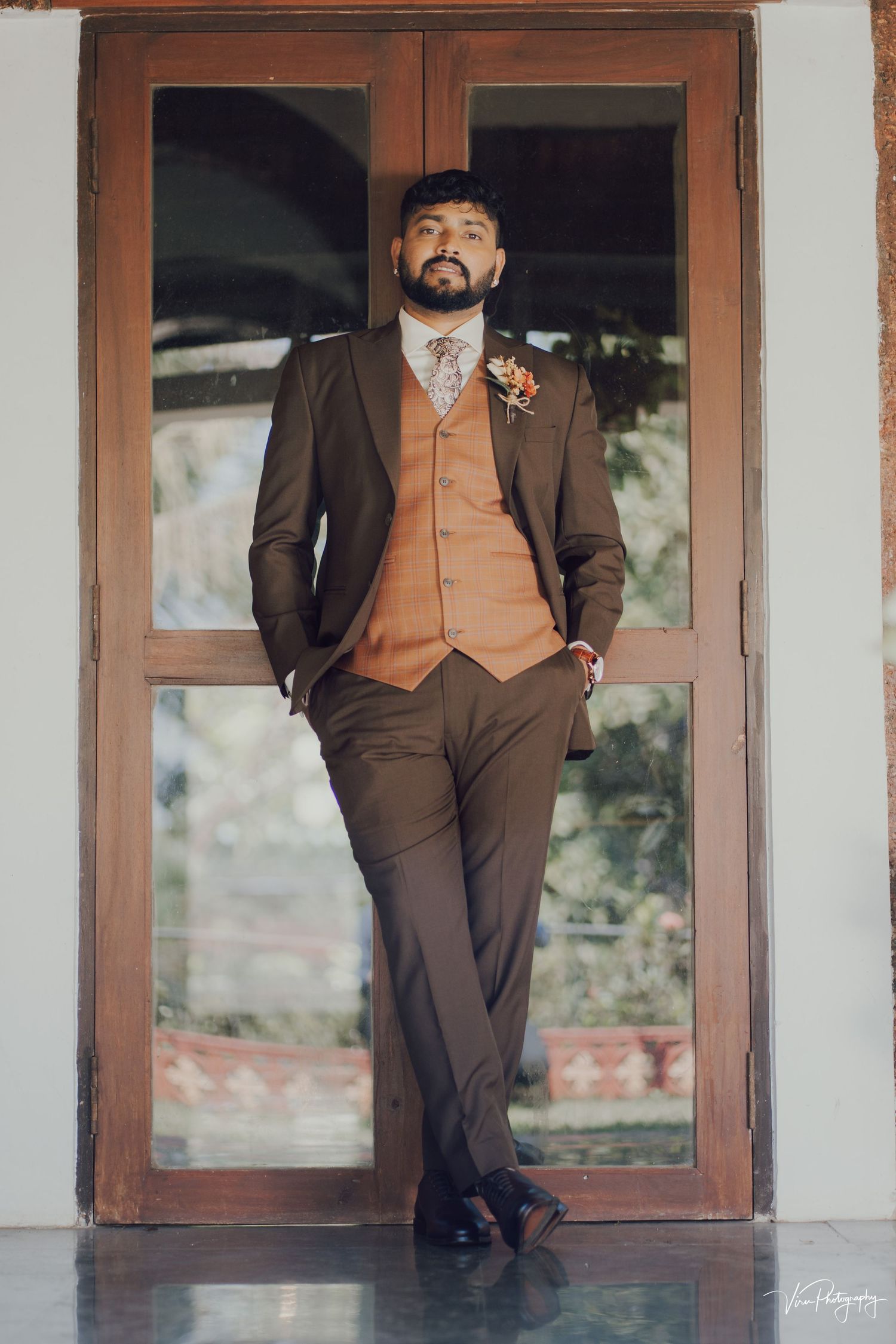 A groom in a brown suit with tan vest poses confidently against wooden doors at a wedding venue.