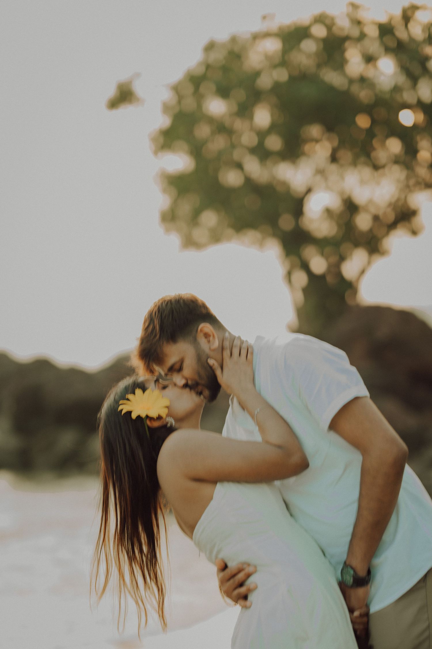 Romantic couple sharing an intimate moment at sunset with bokeh tree background and warm golden lighting.