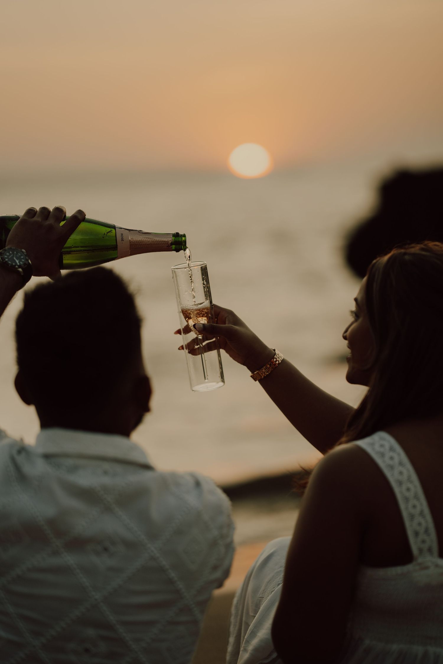 Silhouettes of people toasting with drinks against a sunset sky during a beach celebration.