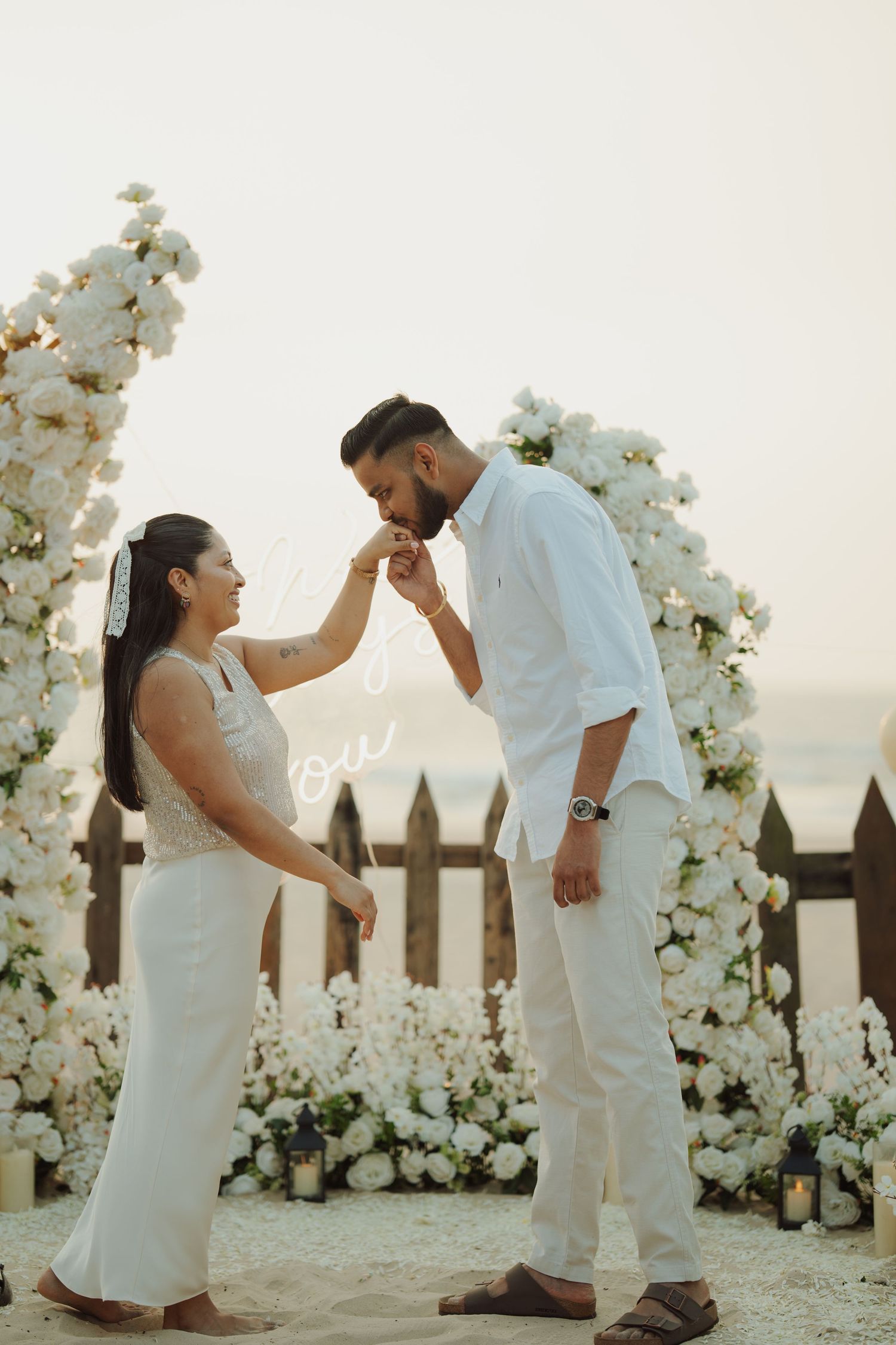 A couple in white attire shares a romantic moment at a floral arch beach proposal setup with white roses.