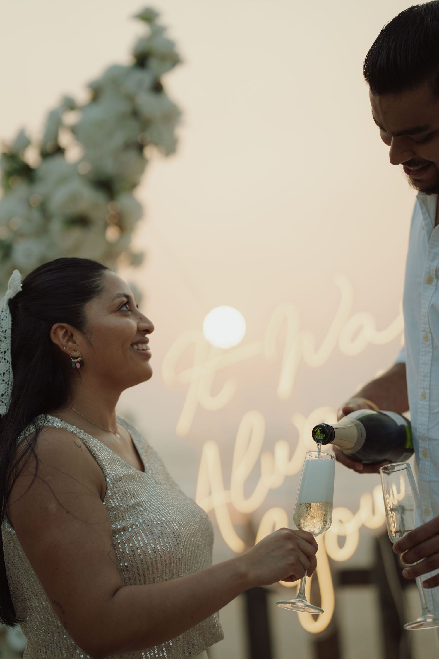 A smiling bride in a sequined dress holds champagne flutes as a man pours bubbly at an elegant wedding celebration.