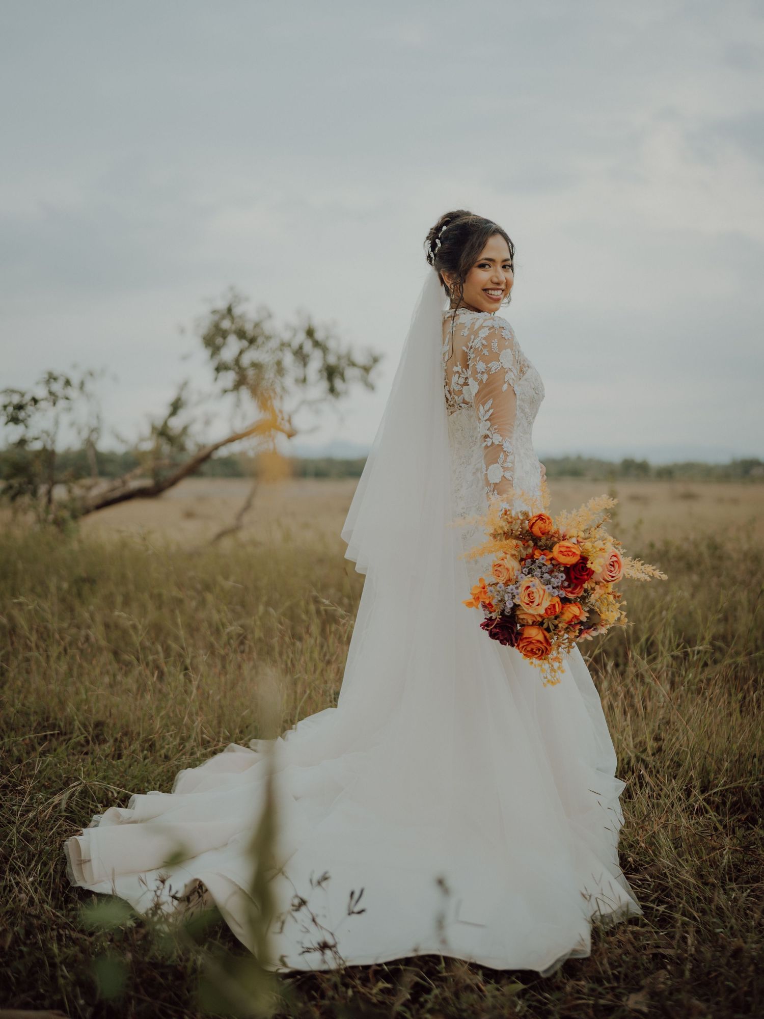 Bride in a lace long-sleeve wedding gown and veil holds an orange floral bouquet while standing in a golden grass field at dusk.