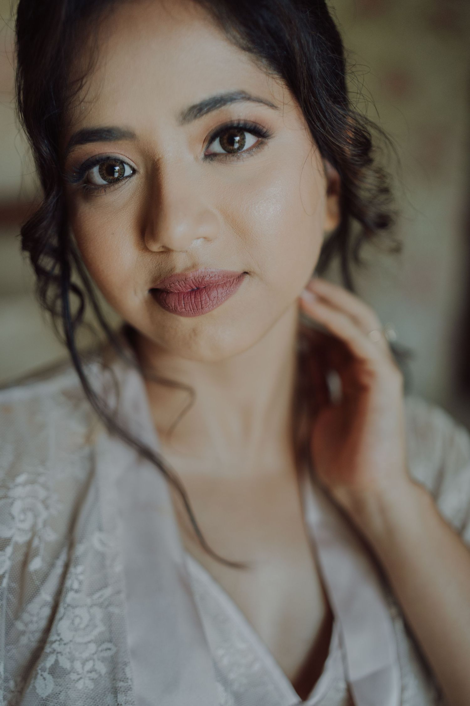 Close-up portrait of a bride with soft wedding makeup and curled tendrils, wearing a white lace robe and touching her neck.