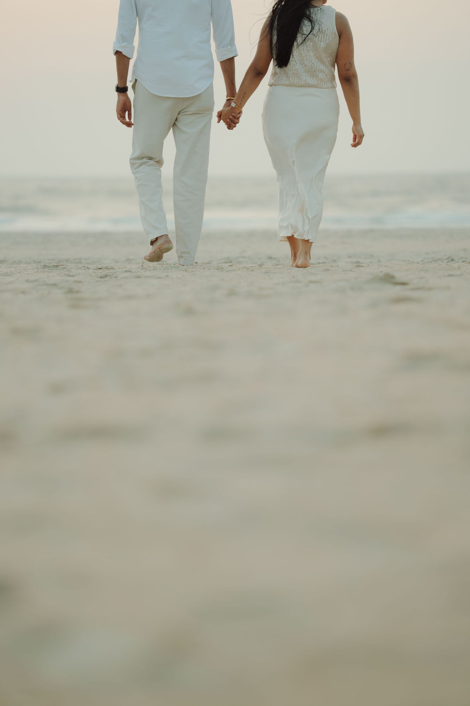 Couple holding hands walking barefoot on sandy beach at sunset wearing white outfits.