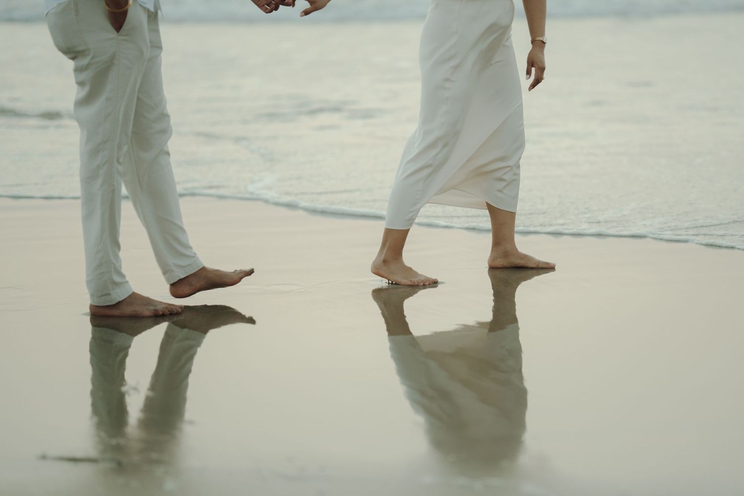 Barefoot couple in white outfits walking on a reflective wet beach shore with calm ocean waves.