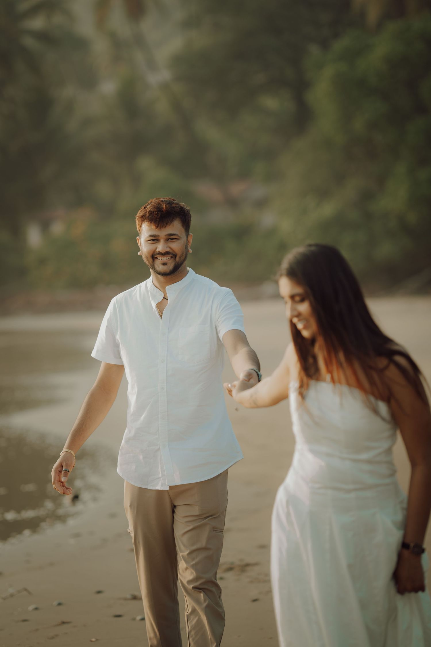 A couple walks together on a sandy beach wearing white and beige casual attire during a romantic moment.