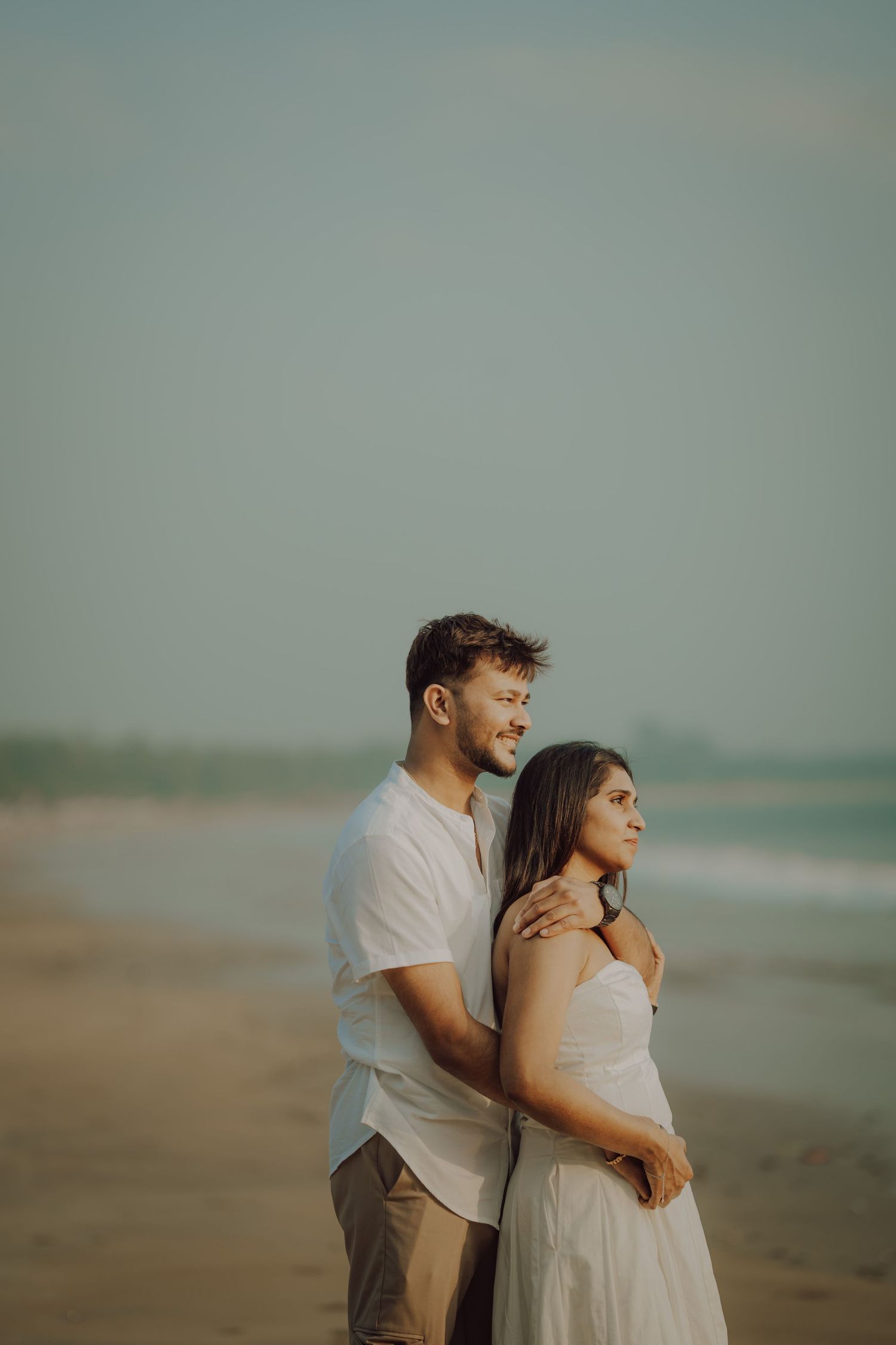 A couple in white outfits embraces while enjoying a romantic moment on a misty beach at sunset.