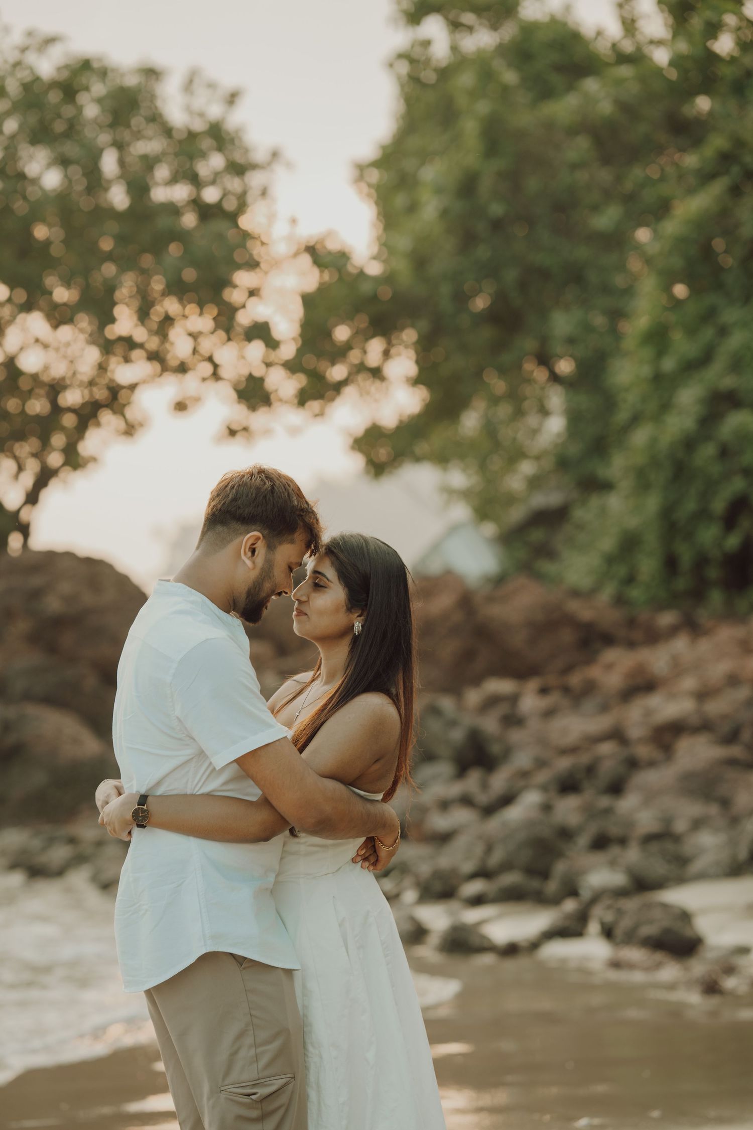 Romantic couple embraces on a rocky beach at sunset while wearing white and beige outfits.