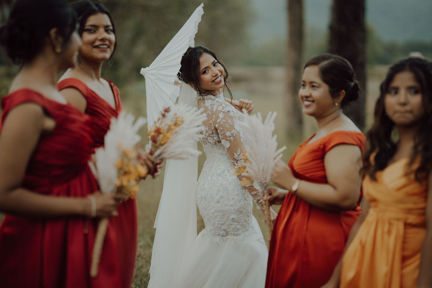 Bride in a lace gown and flowing veil laughs outdoors surrounded by bridesmaids in red and orange dresses holding feather bouquets.