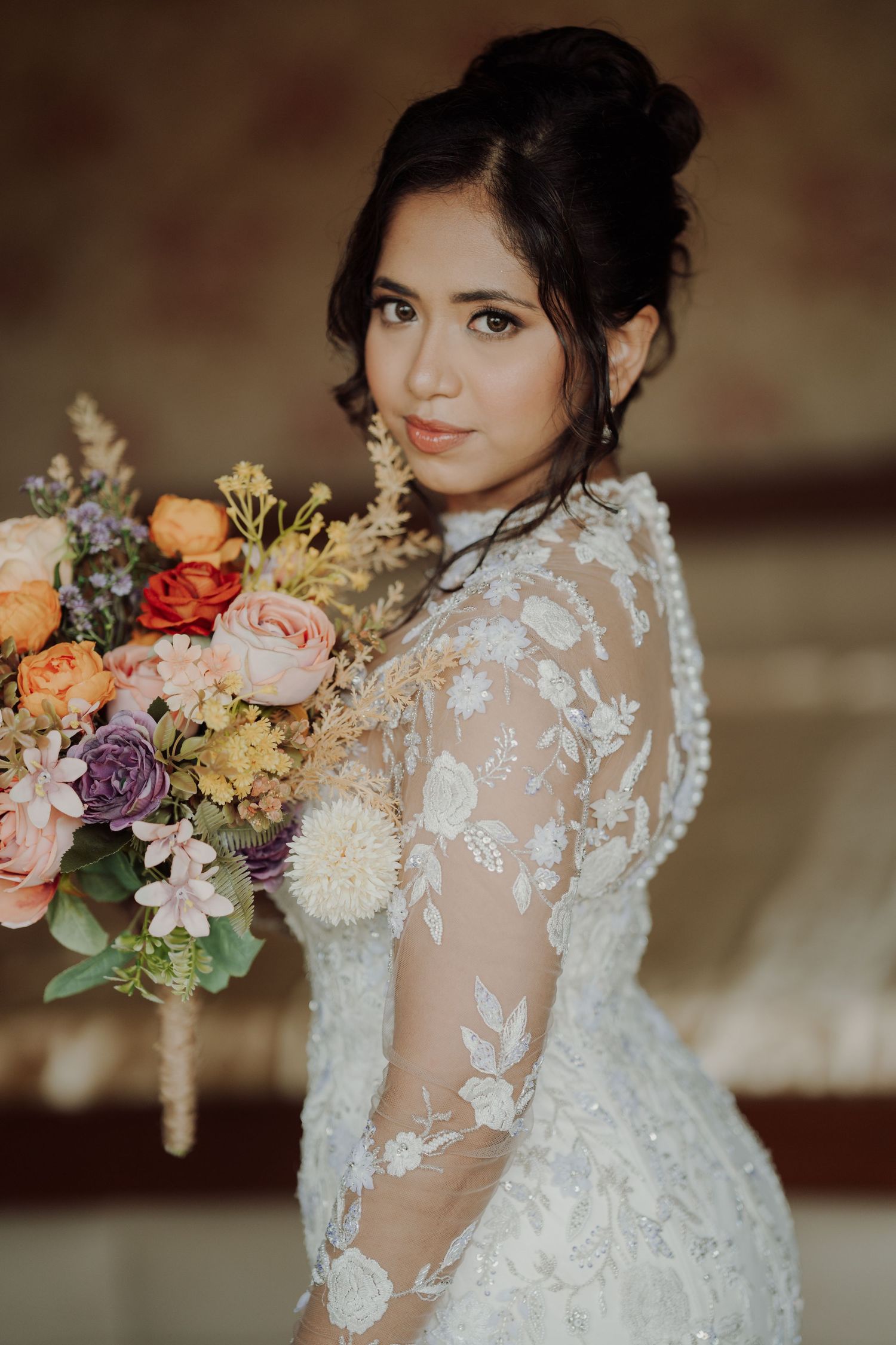 Bride in an embroidered white lace gown holds a colorful bouquet of roses and wildflowers while posing with an elegant updo.