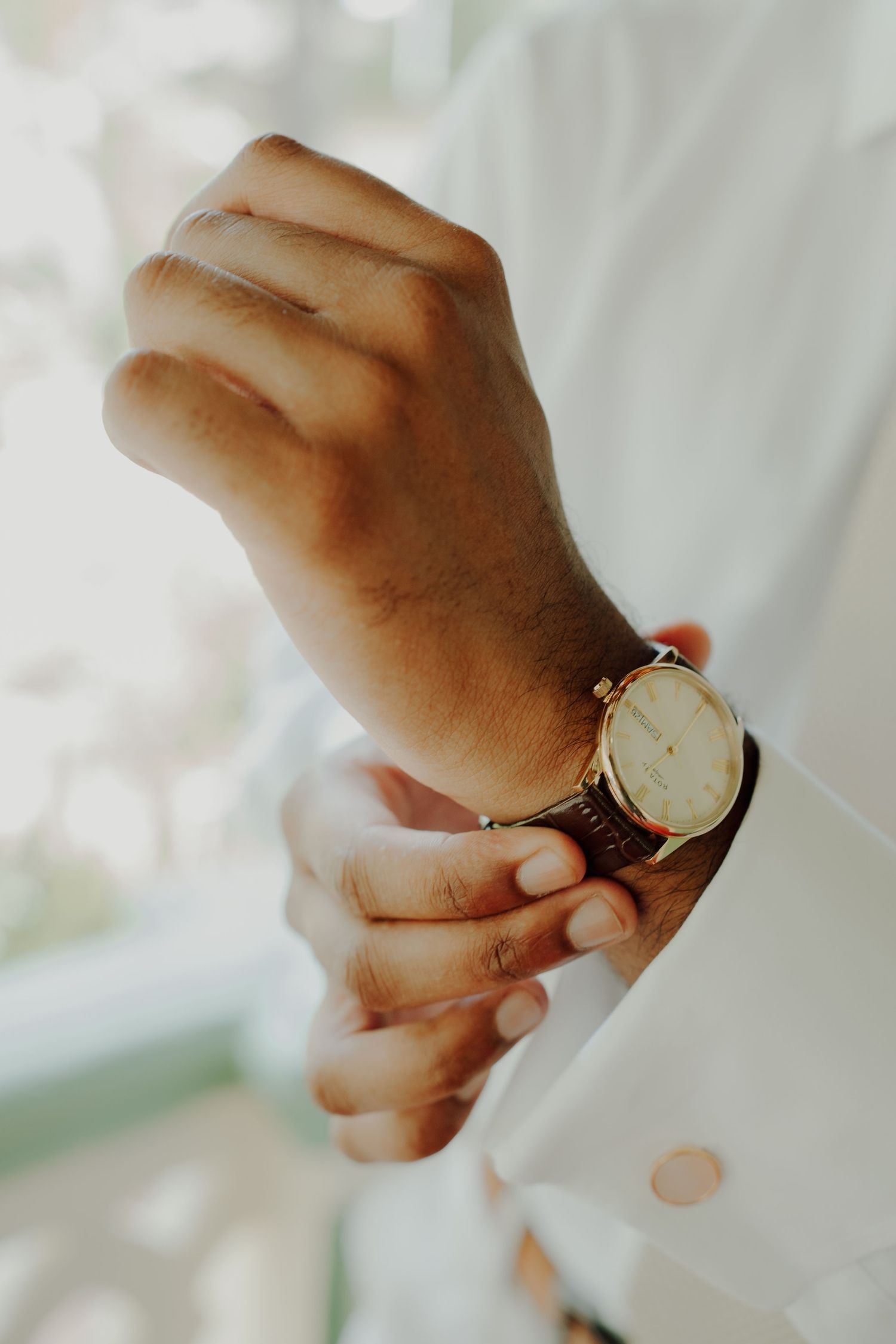 Close-up of a groom adjusting his white shirt cuff and gold cufflink while wearing a gold-faced wristwatch with a brown leather strap.