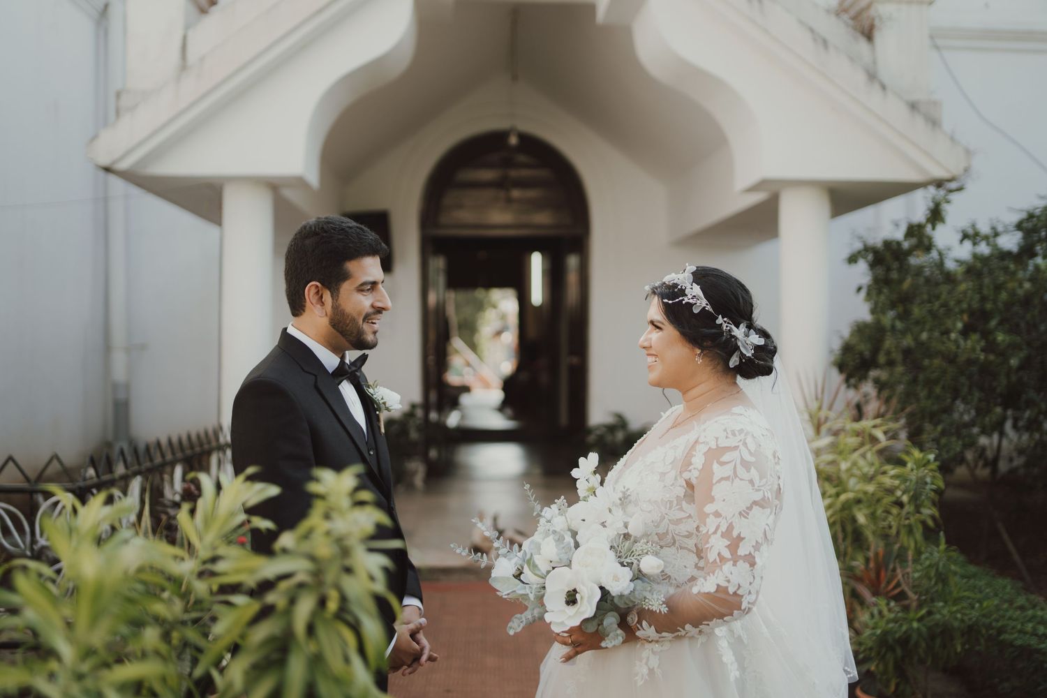 A couple shares an intimate moment surrounded by greenery outside a white church entrance.