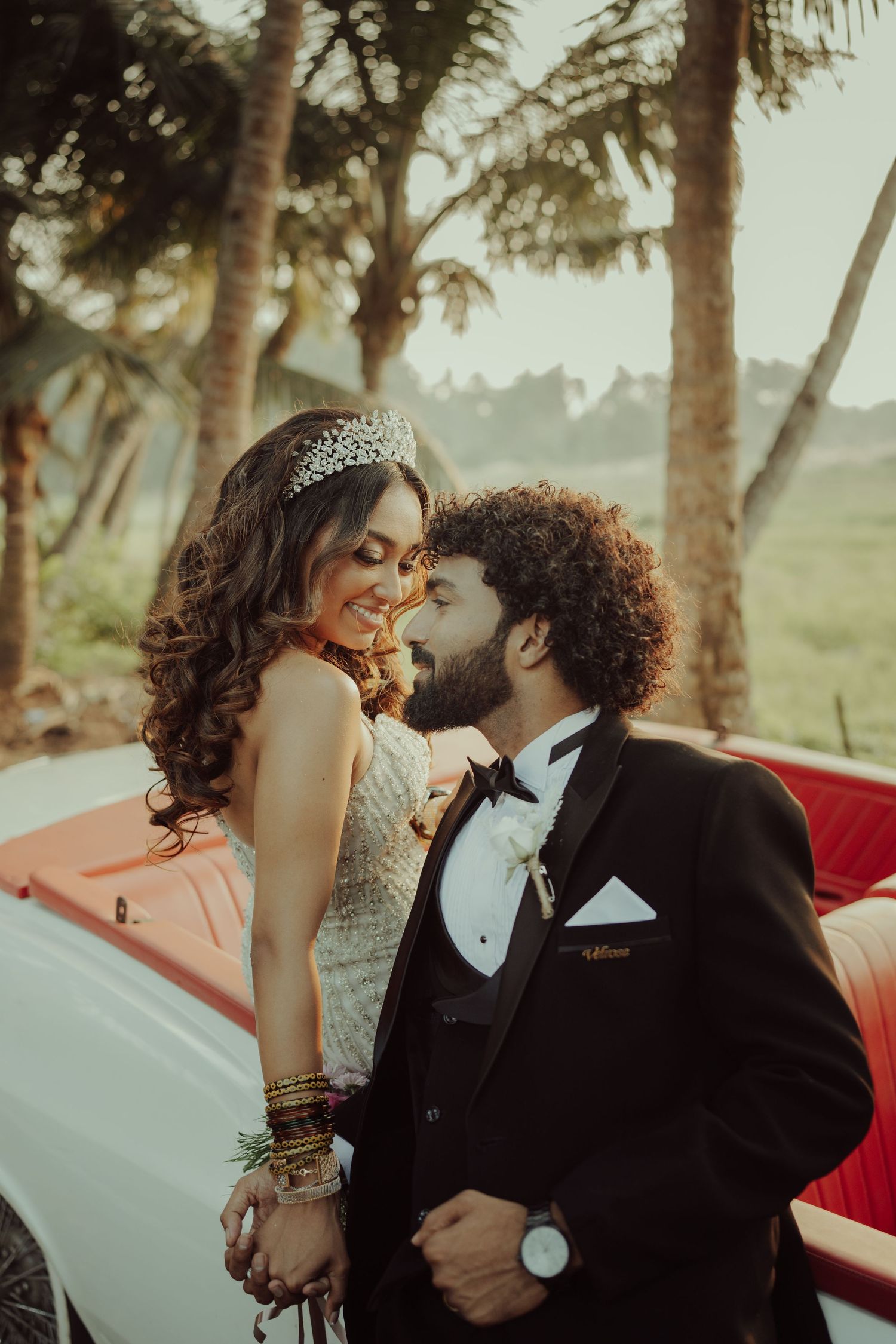 Romantic wedding couple poses by vintage car with palm trees in background.