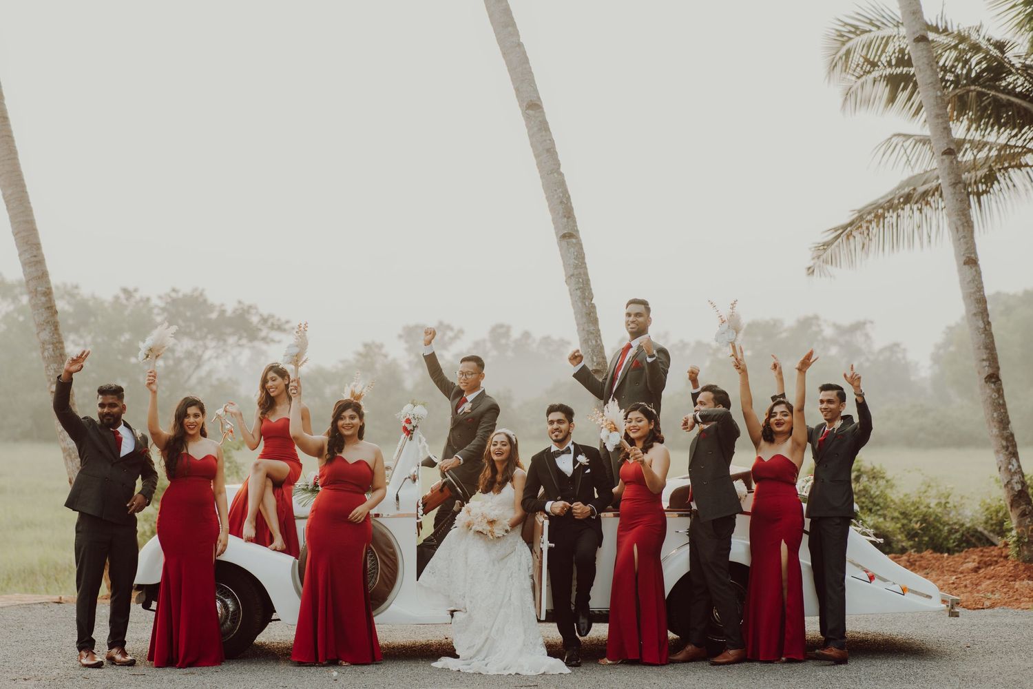 Wedding party in red and black formal attire pose together outdoors with palm trees in a misty background.