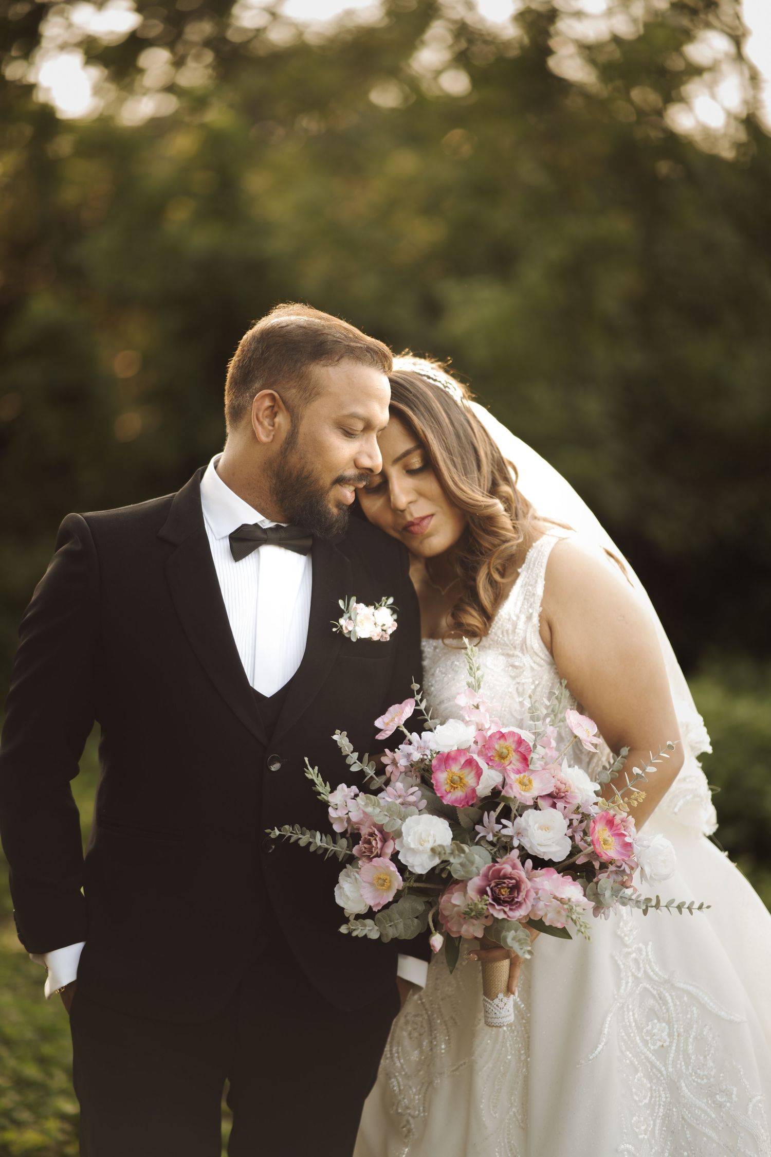 A romantic wedding couple shares an intimate moment outdoors while holding a pink and white floral bouquet.