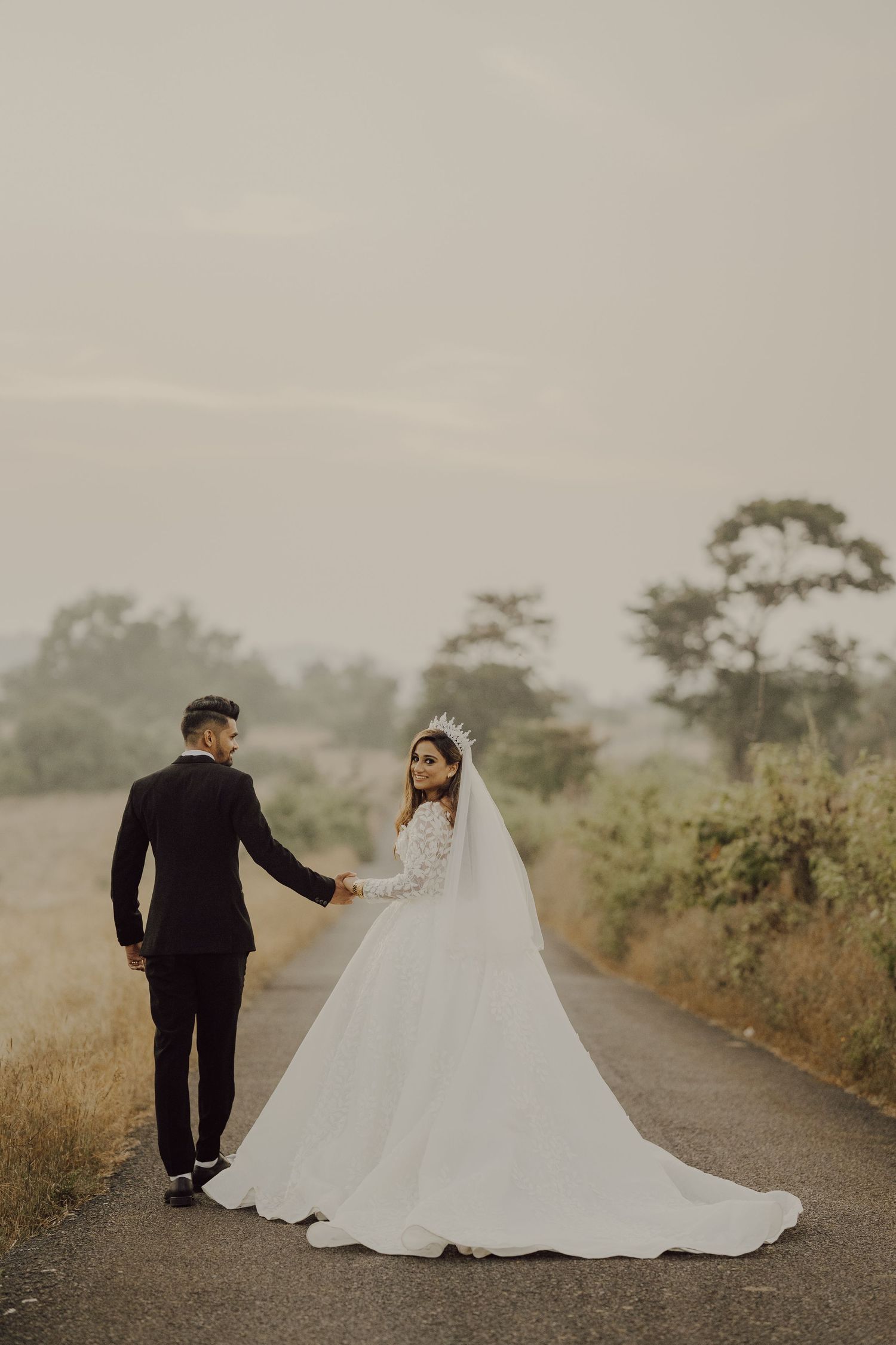 A couple in wedding attire walks down a rural road at dusk with a long white veil trailing behind.