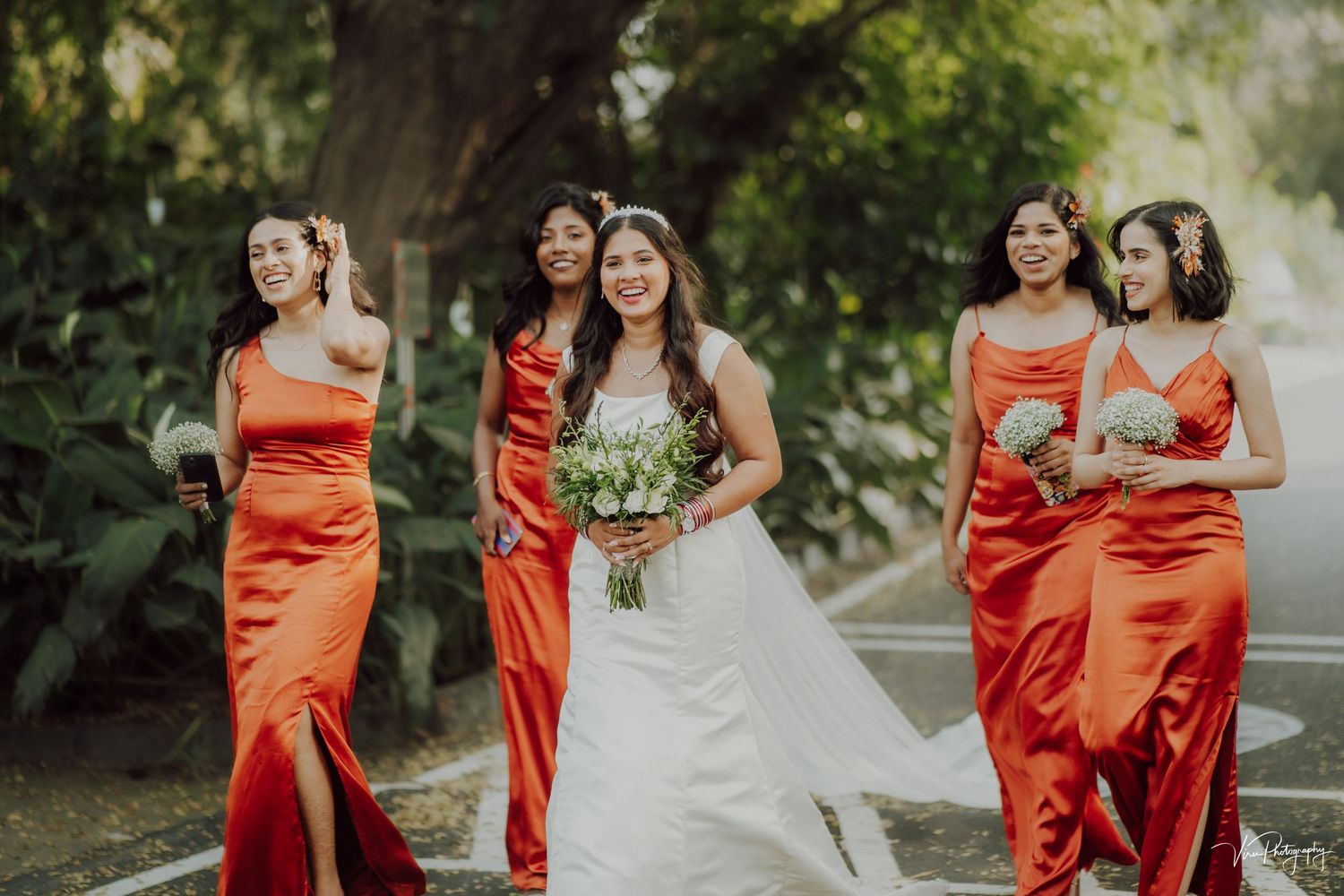 Bridal party walks outdoors wearing vibrant coral satin dresses with white bouquet at garden wedding ceremony.