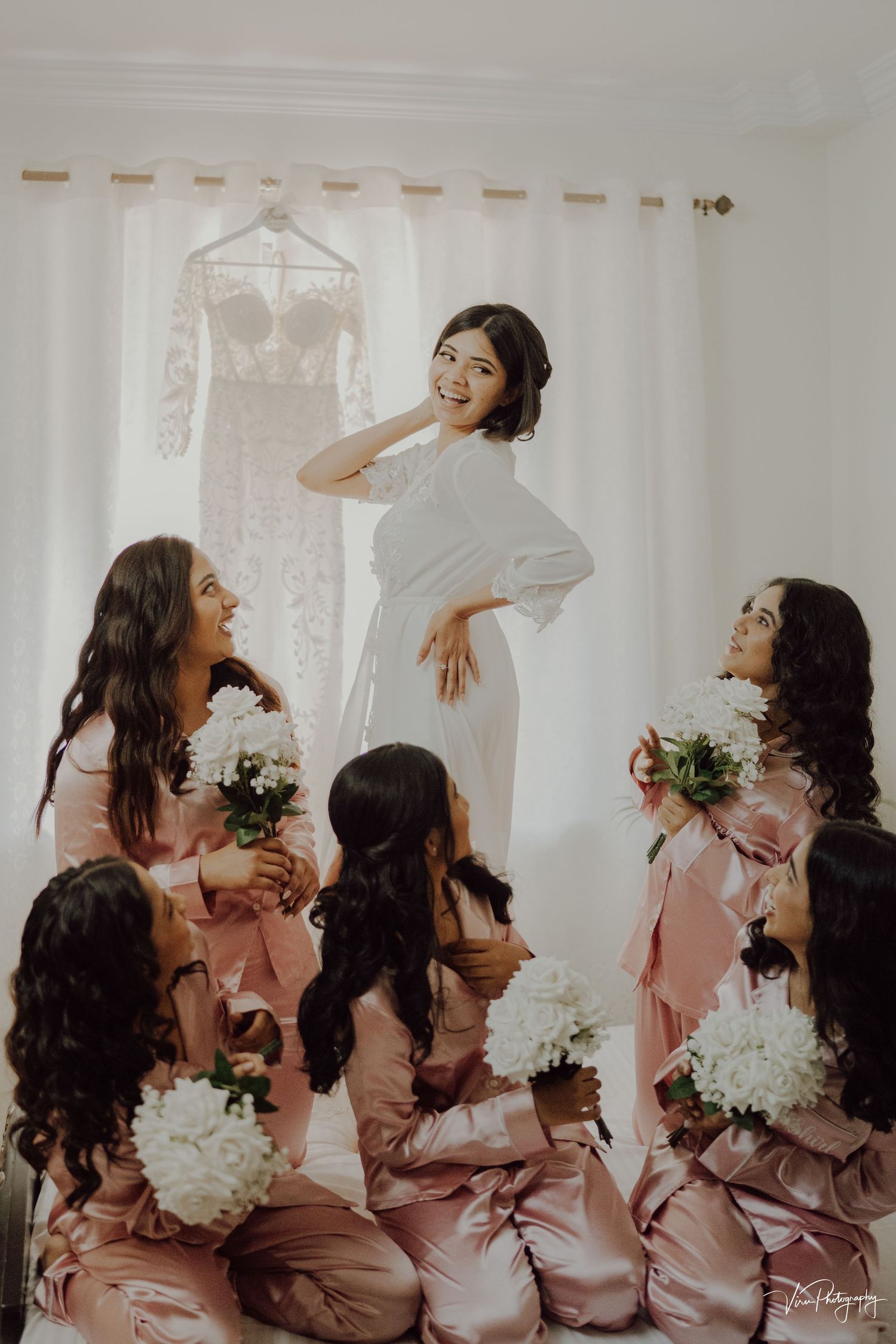 Bridesmaids in pink satin robes gather joyfully around a bride in white during wedding day preparations.