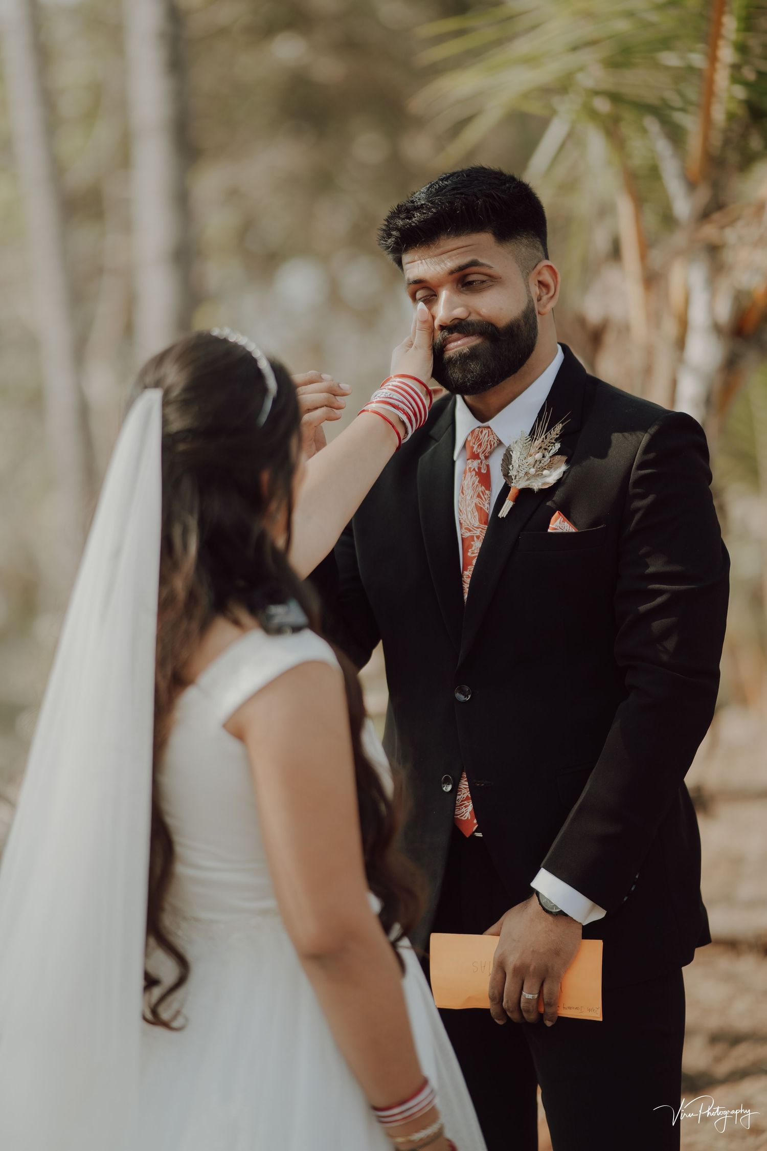 A groom in a black suit and floral tie shows emotion during an intimate outdoor wedding ceremony.