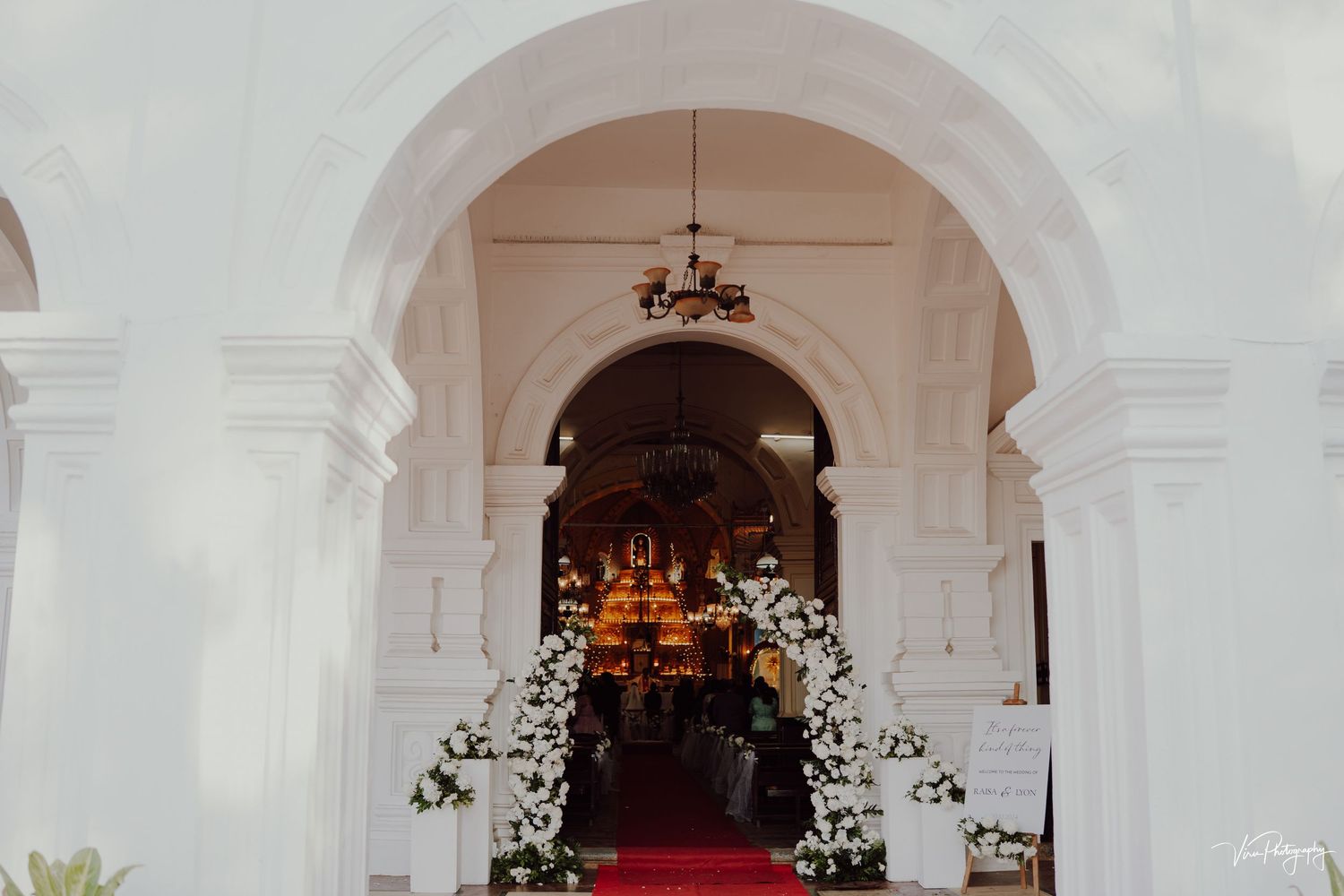 White arched church entrance decorated with floral arrangements and a red carpet leading inside for a wedding ceremony.