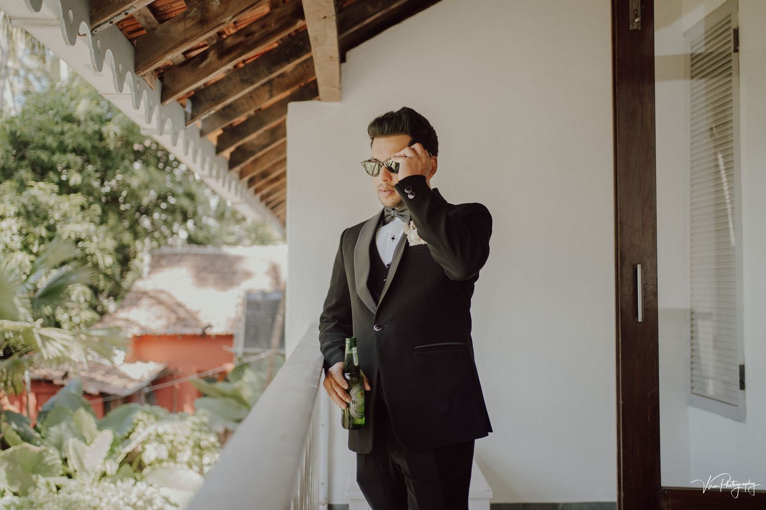 A person in a classic black suit stands outside on a patio, holding a bouquet of flowers near white walls.