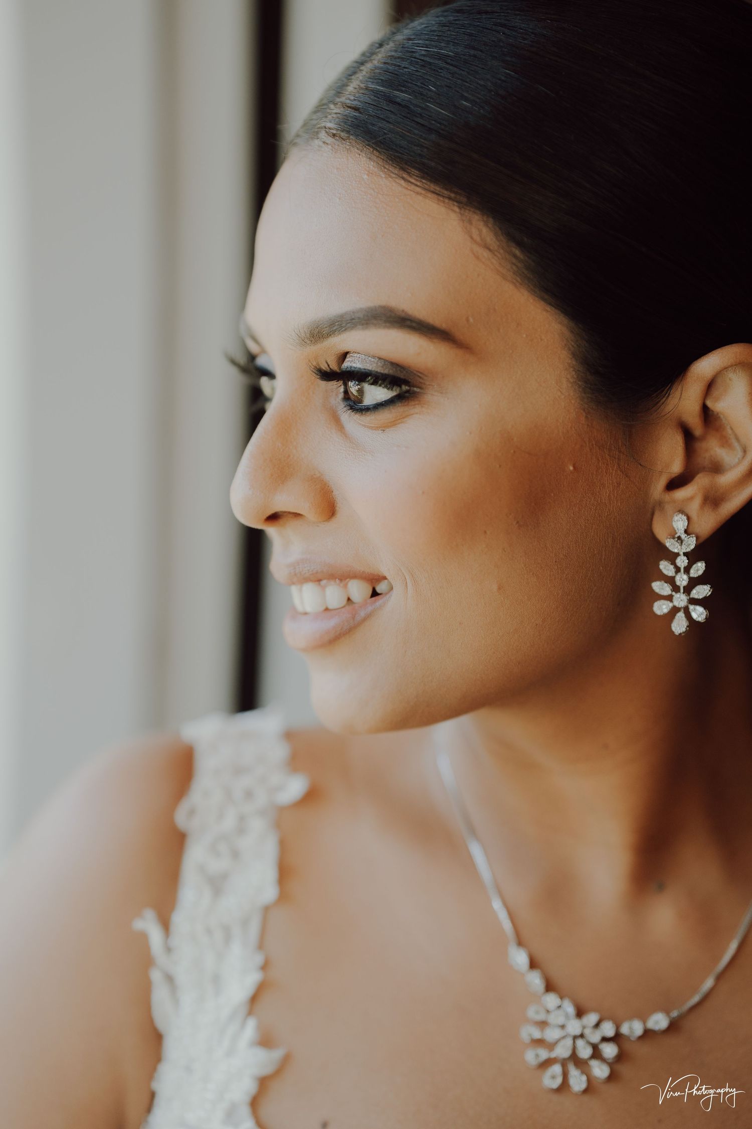 Side profile of a smiling bride wearing crystal floral jewelry and white lace wedding dress near a window.
