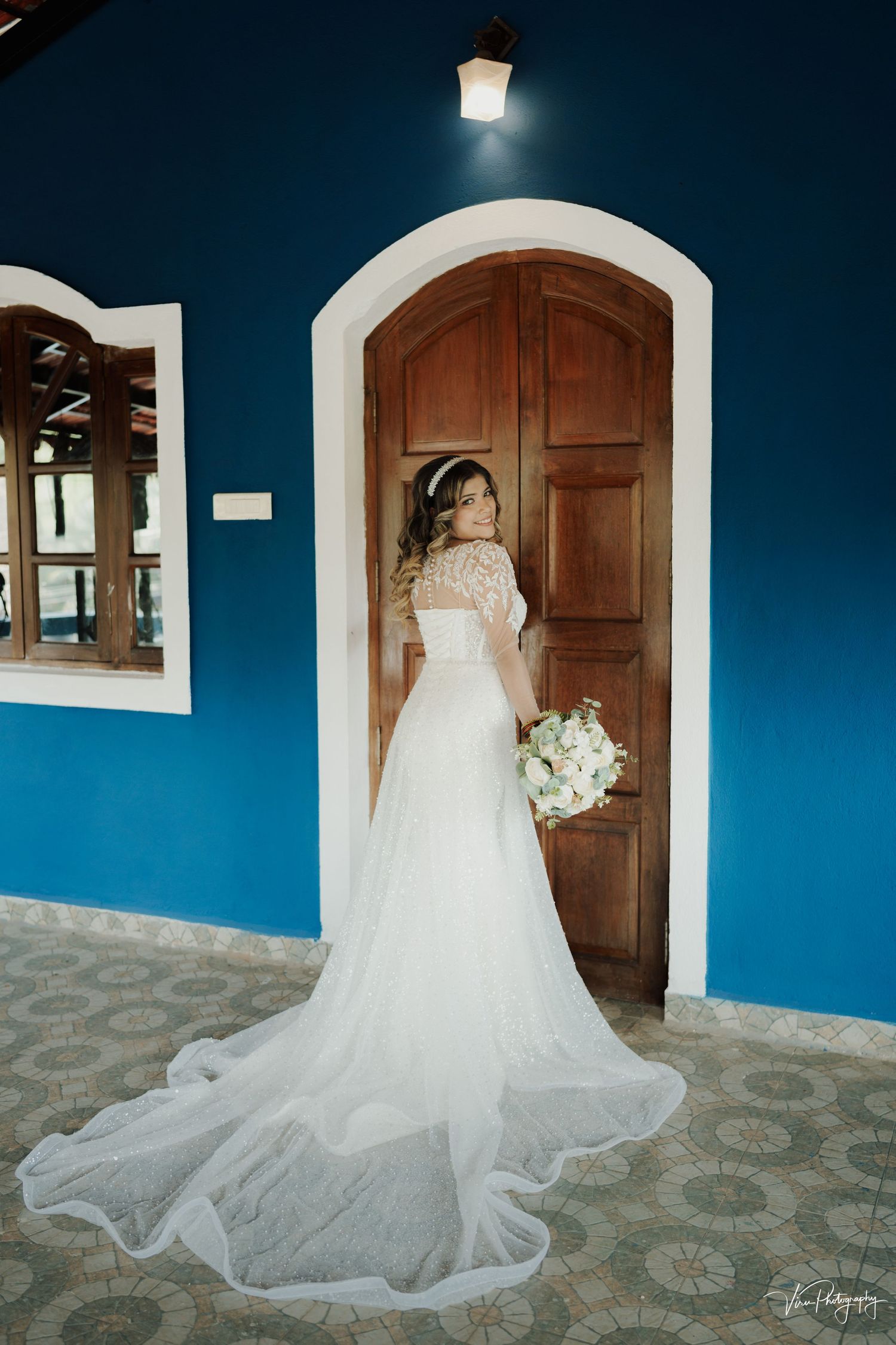 A bride in a flowing white lace gown stands before a wooden door against a vibrant blue wall with a bouquet.