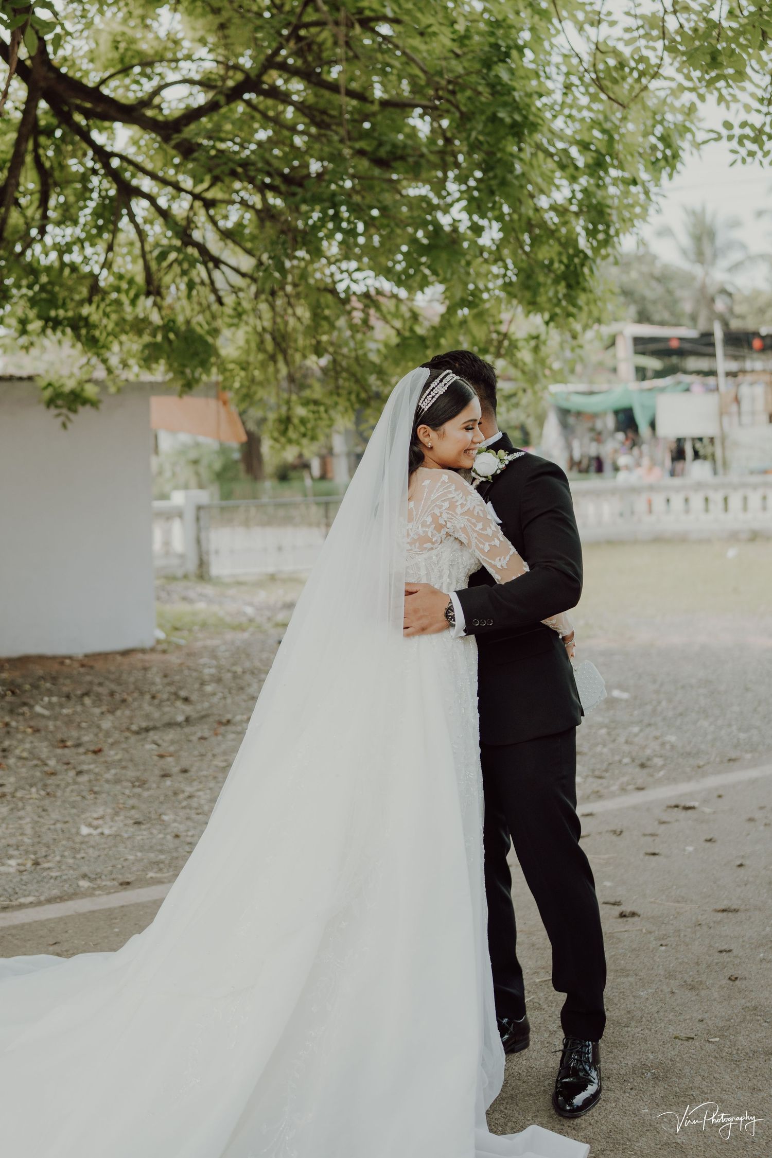 A romantic wedding moment captured outdoors with a long flowing white veil and lace dress under green tree branches.