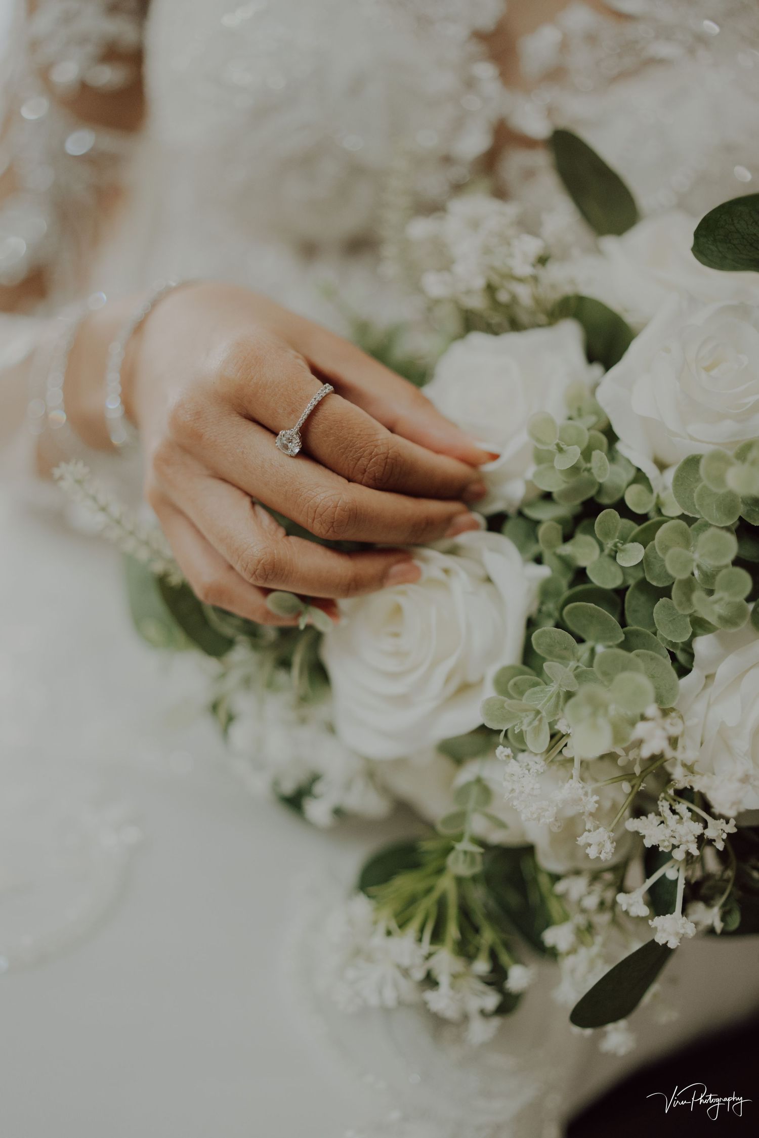A delicate hand with an engagement ring gently touches a bridal bouquet of white roses and green hydrangeas.