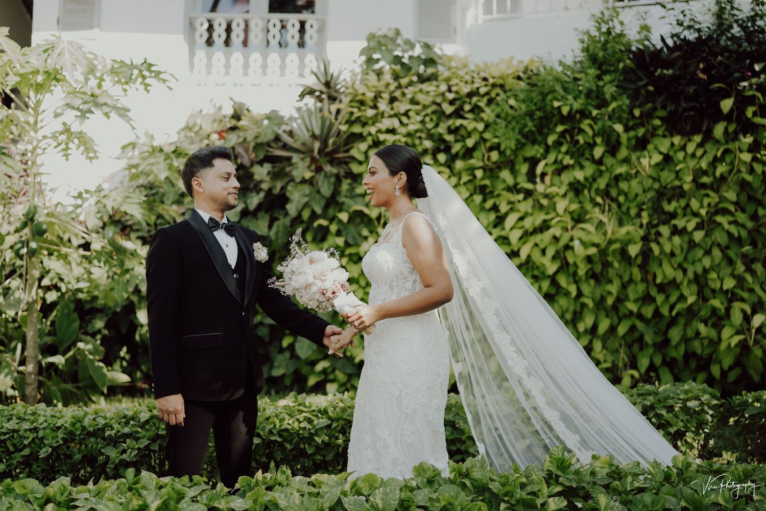 A formal wedding portrait taken in a lush garden with greenery and white architecture in the background.