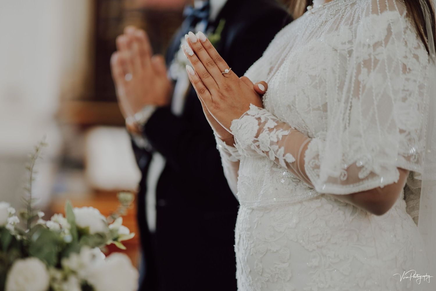 Hands clasped in prayer at a wedding ceremony with white lace sleeves and floral decorations visible in the background.