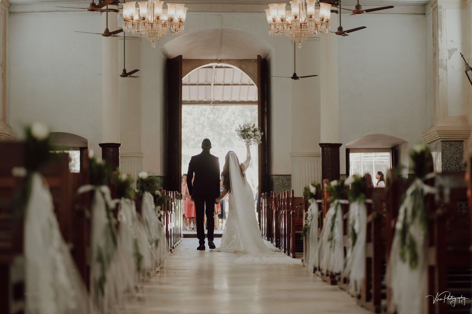 Two silhouettes walk down a church aisle decorated with floral arrangements and illuminated by elegant chandeliers.